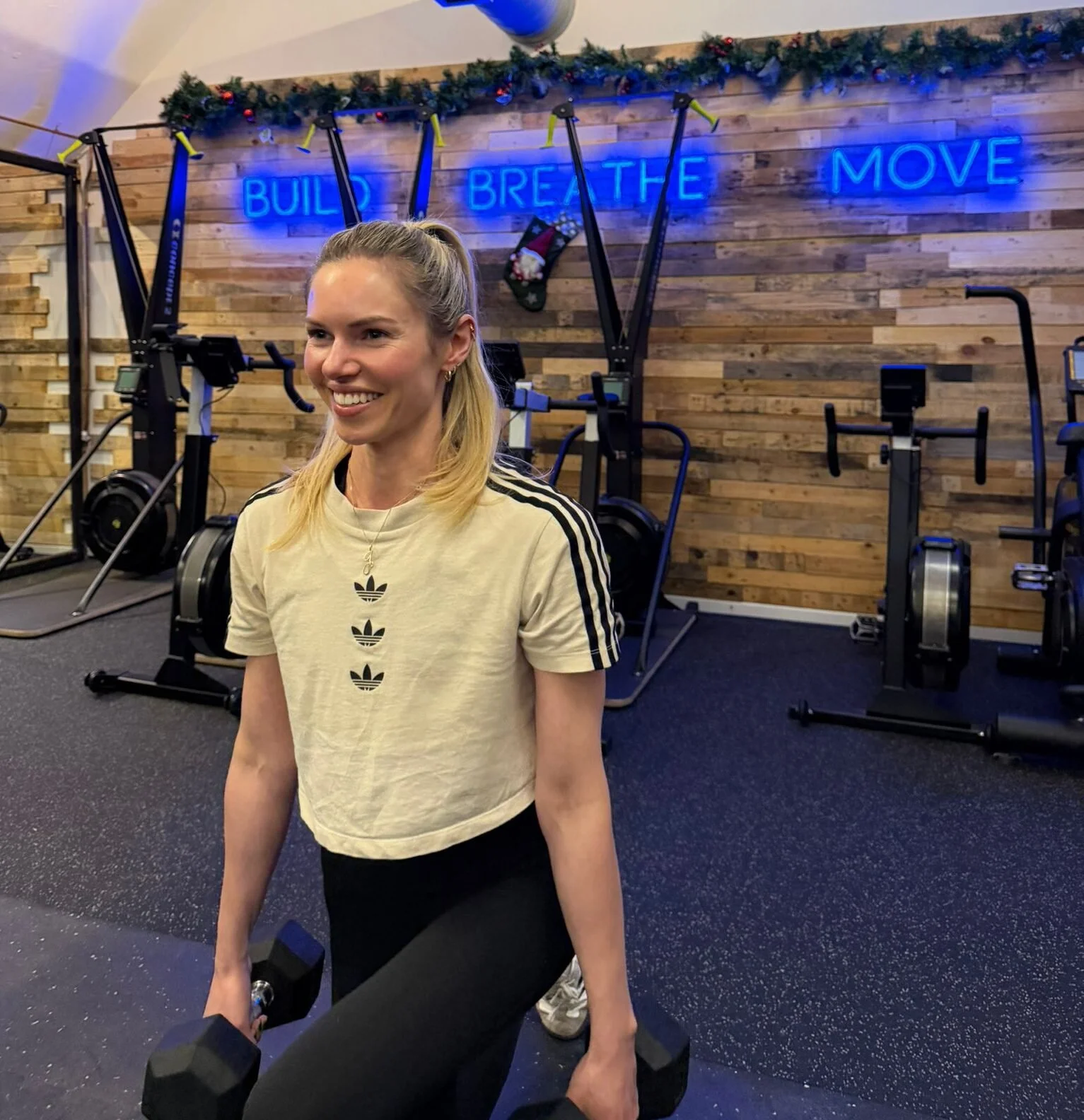 A woman with blonde hair in a ponytail, smiling and holding black dumbbells in a gym. The background features wooden panel walls, gym equipment, and a neon sign with the words 'BUILD BREATHE MOVE' in blue, with holiday decorations hanging above.
