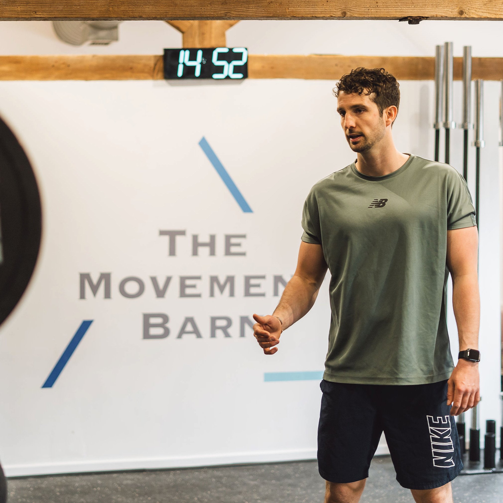 A woman with short blonde hair, wearing a sleeveless rust-colored top and beige pants, sitting on a beige bench, writing in a notebook. Behind her is a large green plant, a wooden slatted wall, and a framed abstract art piece. Sunlight is streaming through a window, casting shadows on the wall.