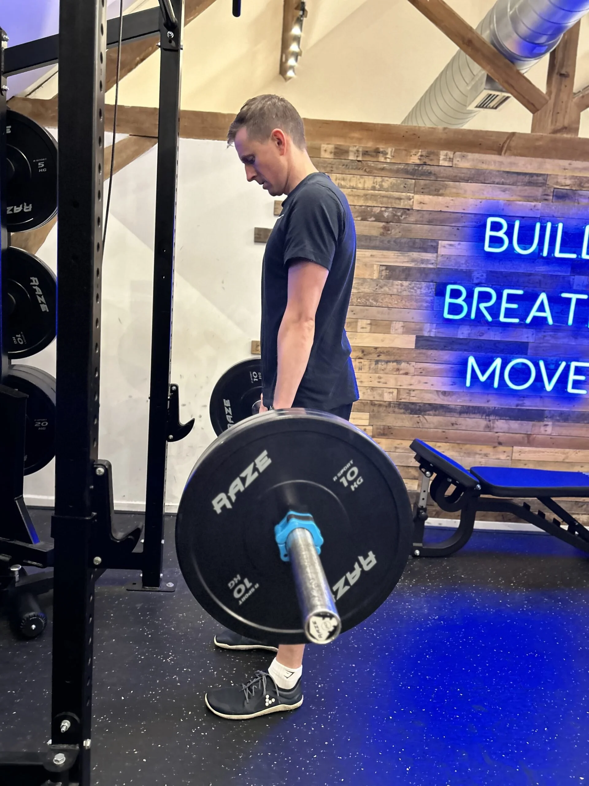 A man working out with a loaded barbell in a gym with wooden walls and neon sign that reads 'Building Breath Moving'.