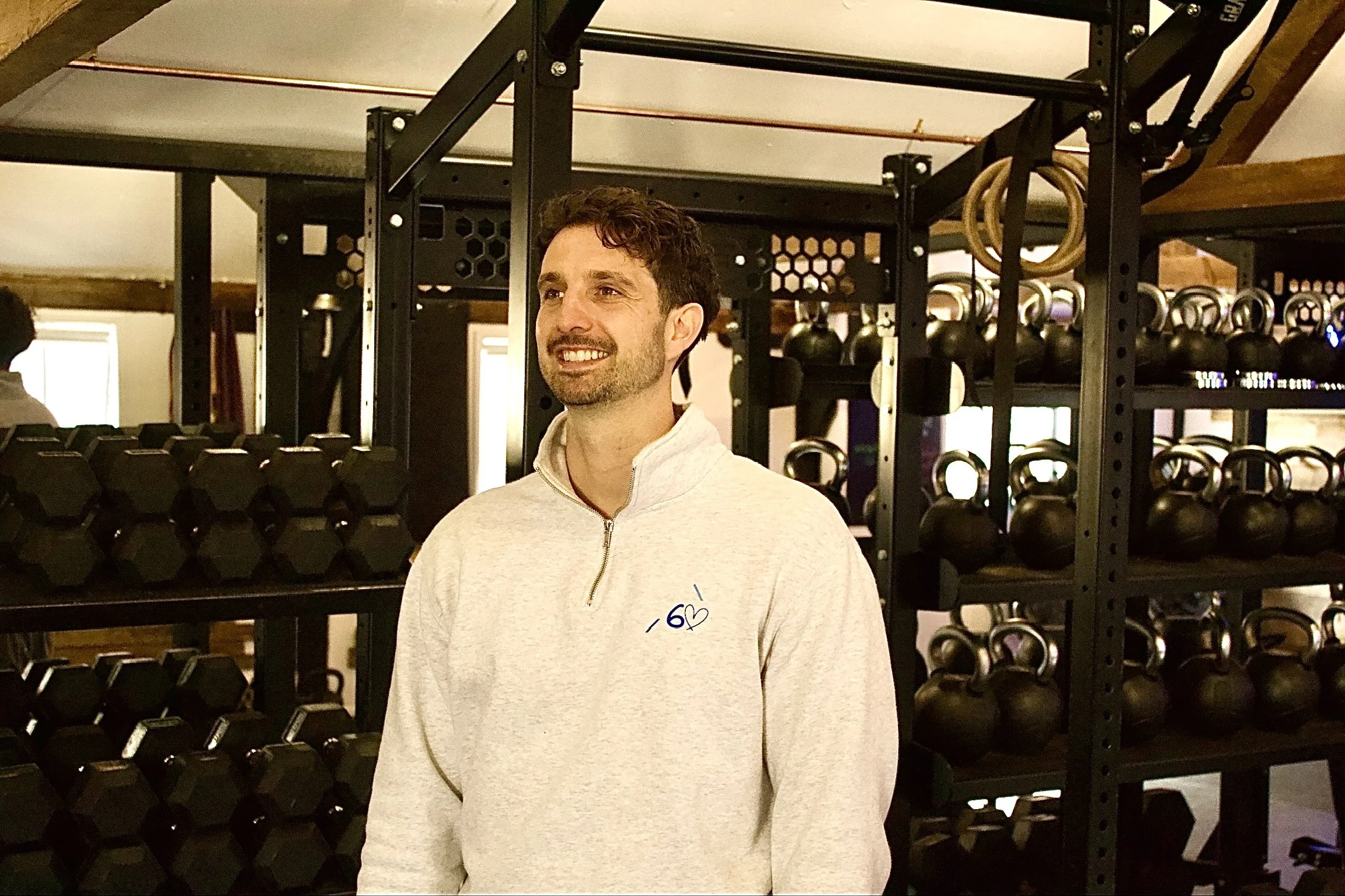 A woman with short blonde hair, wearing a sleeveless rust-colored top and beige pants, sitting on a beige bench, writing in a notebook. Behind her is a large green plant, a wooden slatted wall, and a framed abstract art piece. Sunlight is streaming through a window, casting shadows on the wall.