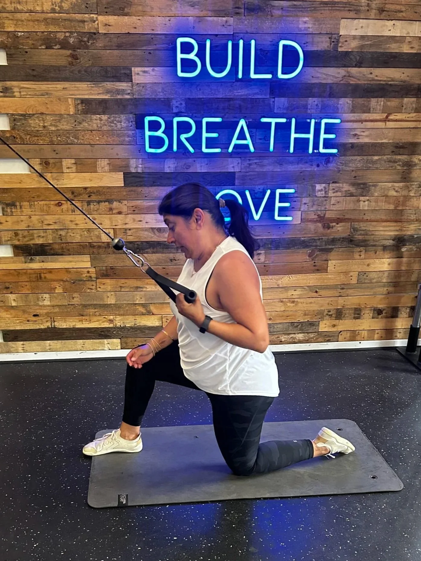 A woman is exercising on a padded mat in a gym, using a cable pulley for a workout. The background features a wooden wall with a neon sign that says 'BUILD BREATHE MOVE' in blue lights.