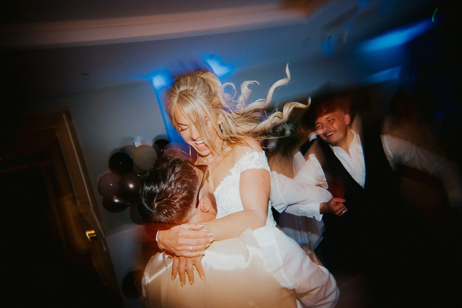 A bride with long blonde hair dancing with a groom during her wedding reception, surrounded by other guests, with balloons in the background.