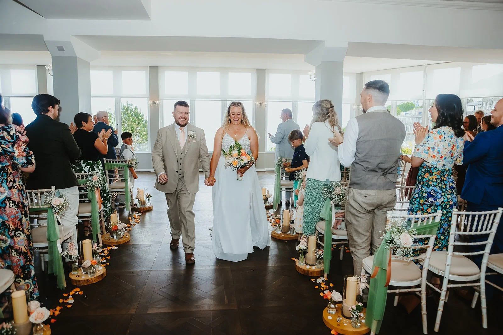 A bride and groom walking hand in hand down the aisle at their wedding ceremony, surrounded by seated and standing guests in a bright, airy room with large windows and floral decorations.