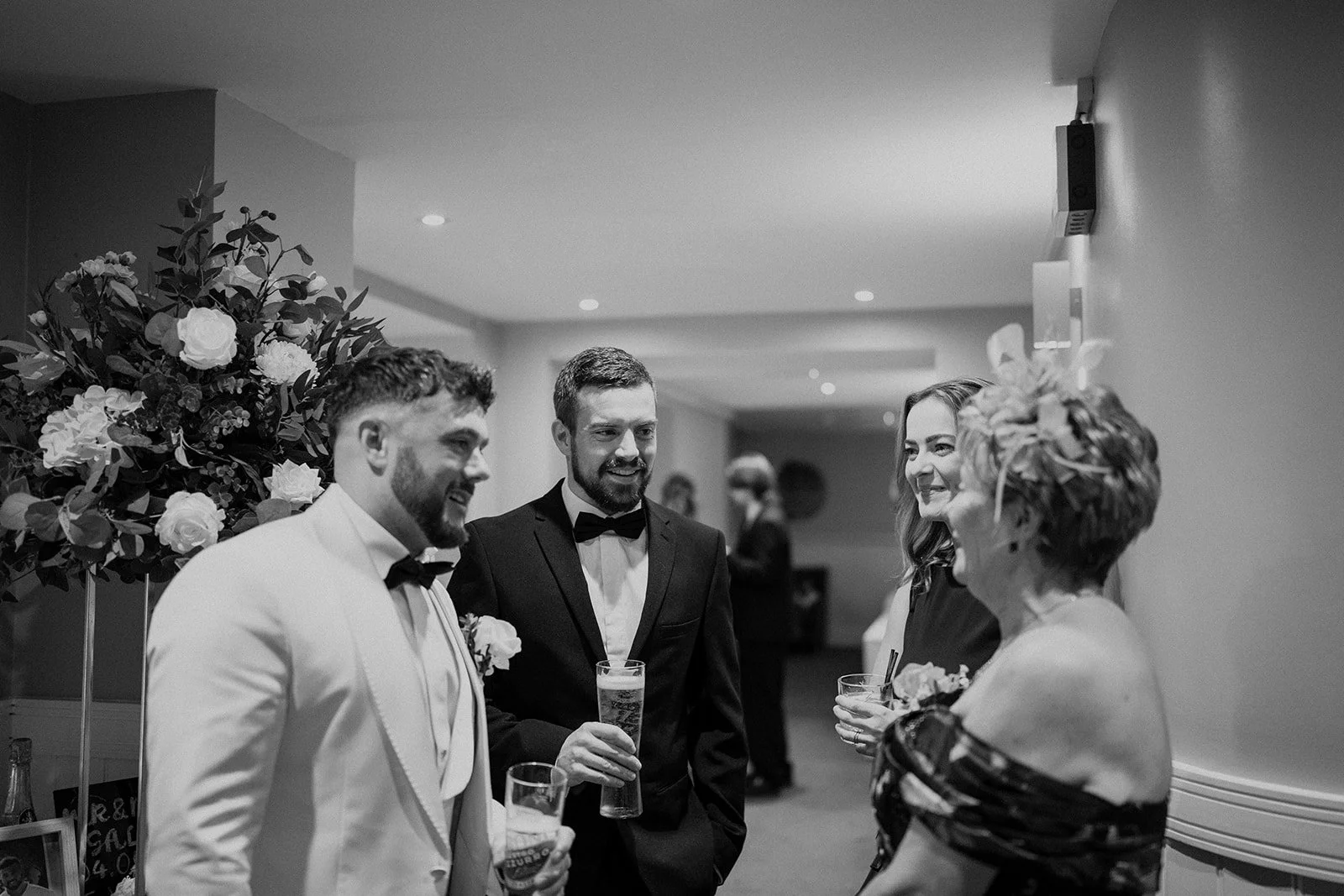Group of four people in formal attire socializing at a celebration, with floral arrangements in the background.