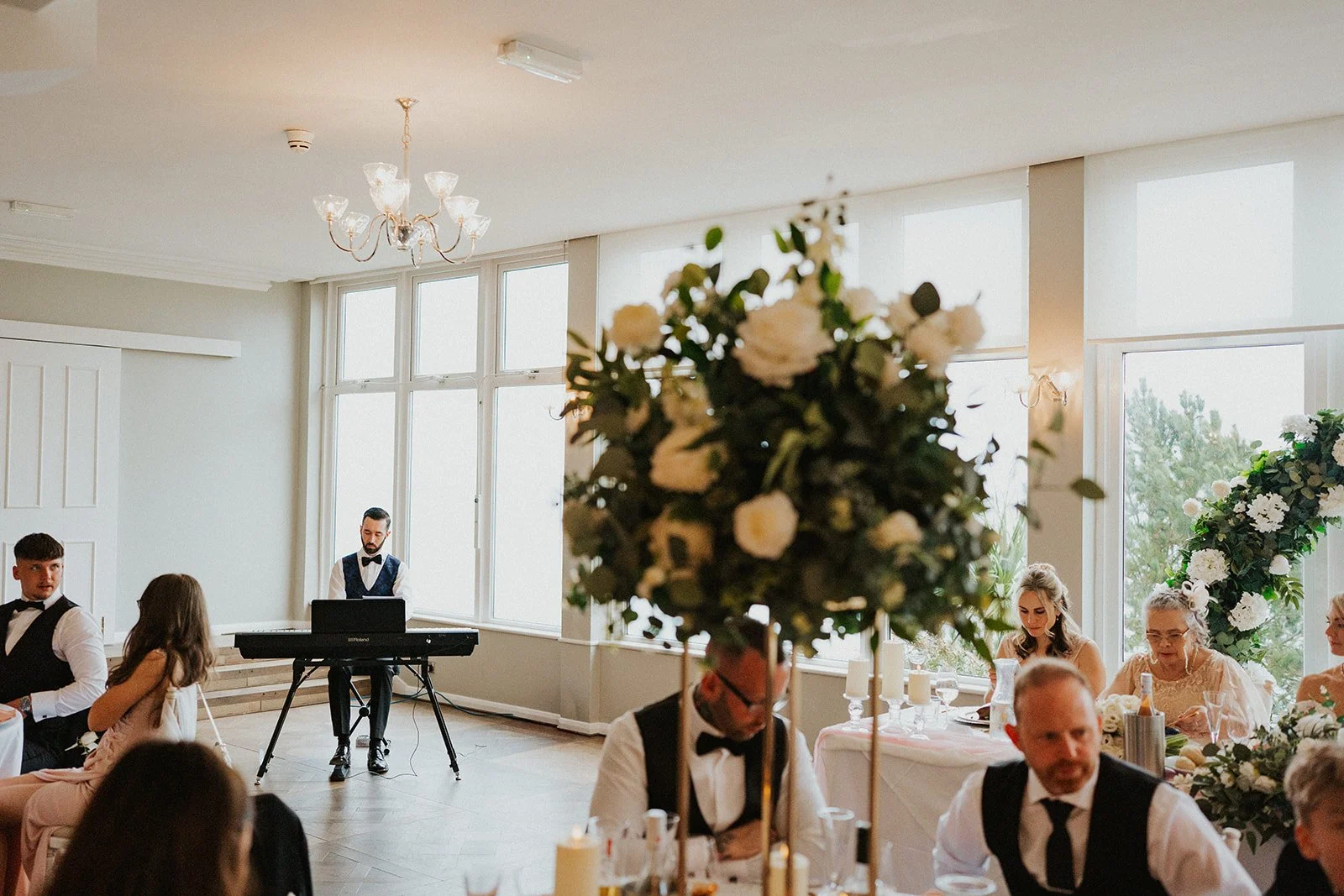Indoor wedding reception with floral centerpieces, guests seated at tables, and a band with a keyboard performing in front of large windows.