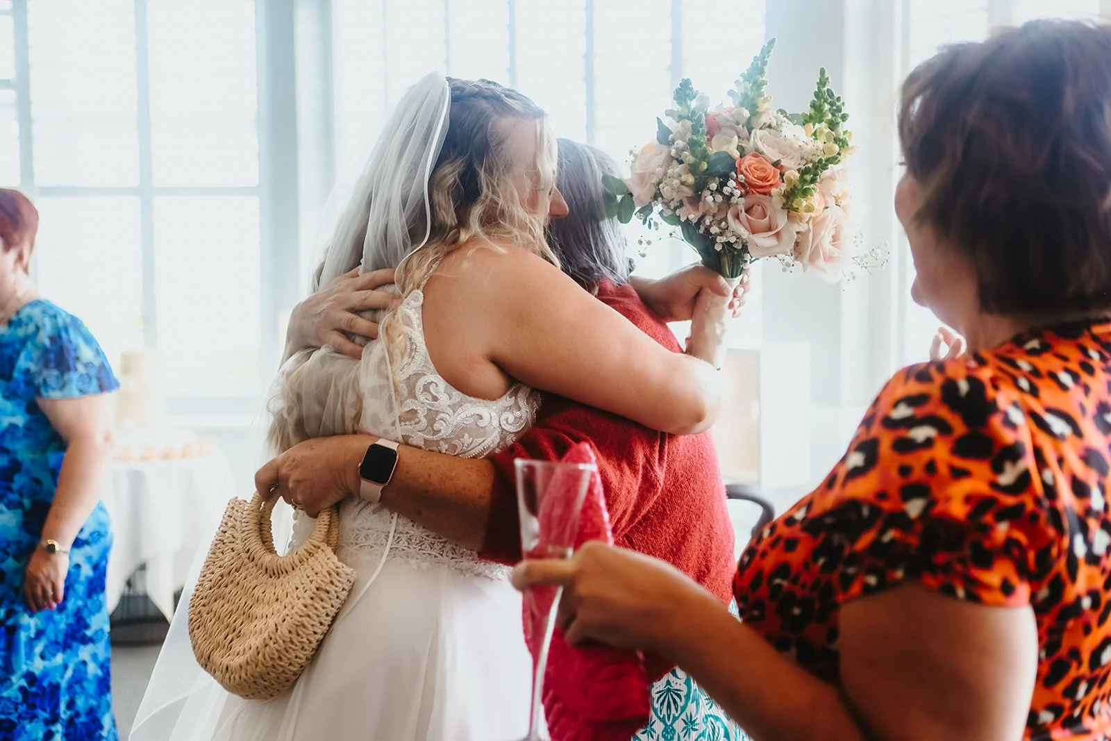 A bride hugging a guest while holding a bouquet, at a wedding reception with other guests in colorful dresses.