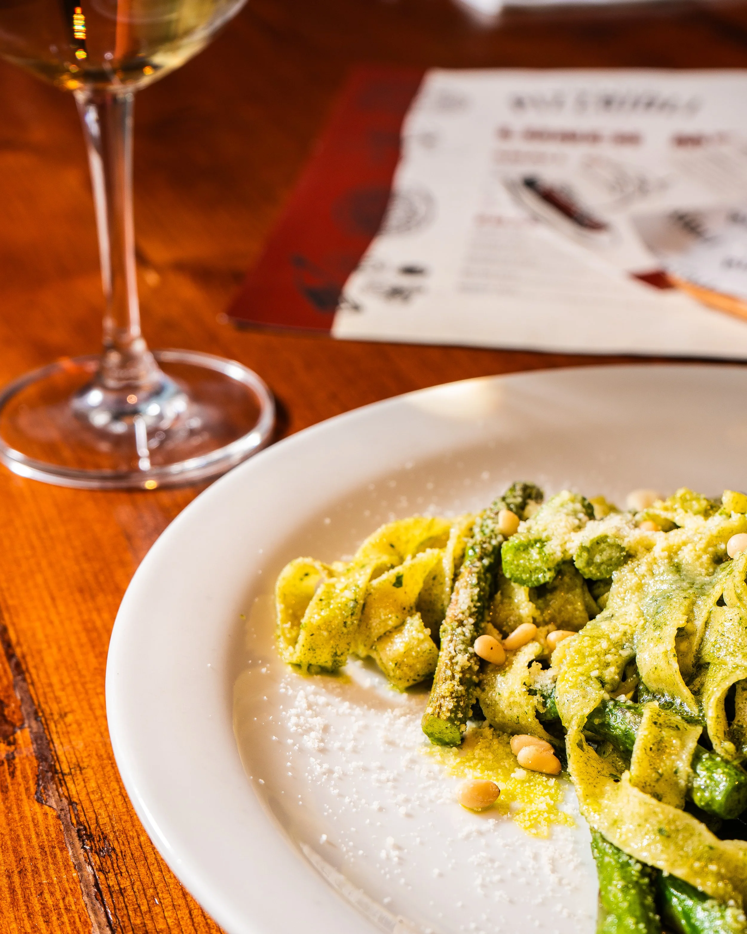 Plate of green pasta with pesto, pine nuts, and grated cheese on a wooden table, with a glass of white wine and restaurant menu in the background.