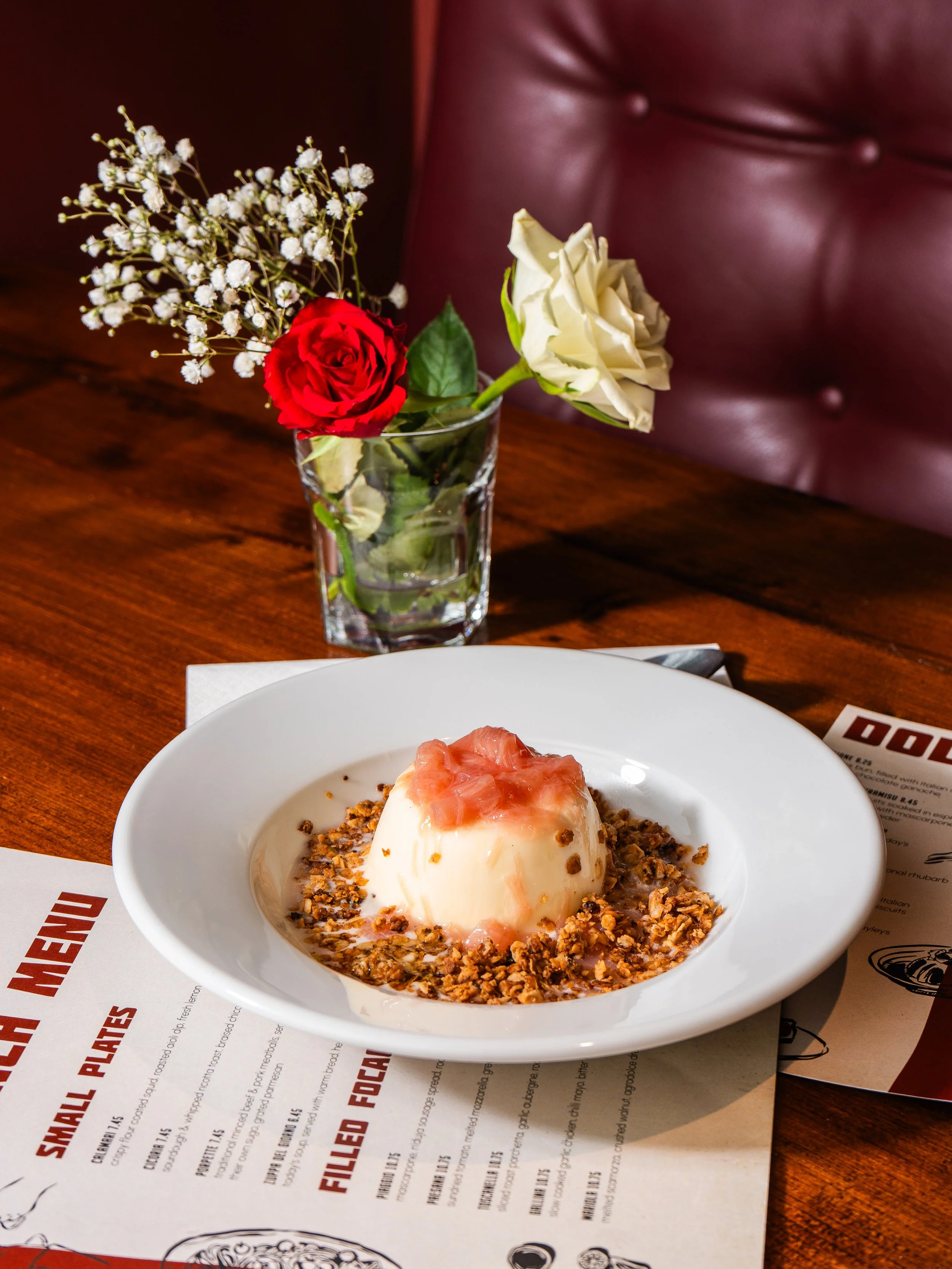 A panna cotta dessert, surrounded by crushed nuts, on a white plate on a wooden table. Behind it, a glass of water holding a single red rose and white flowers. A printed Pizzaioli Ayr menu is also on the table.