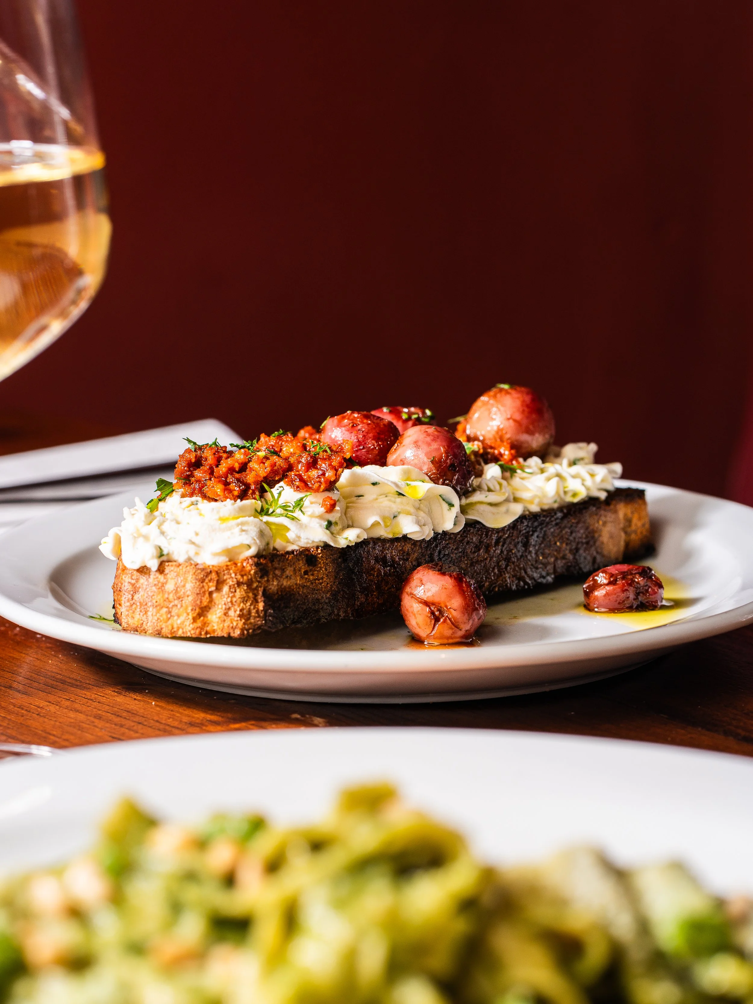 A plate with a slice of toasted sourdough bread topped with whipped ricotta, roasted grapes, and a glass of white wine nearby.
