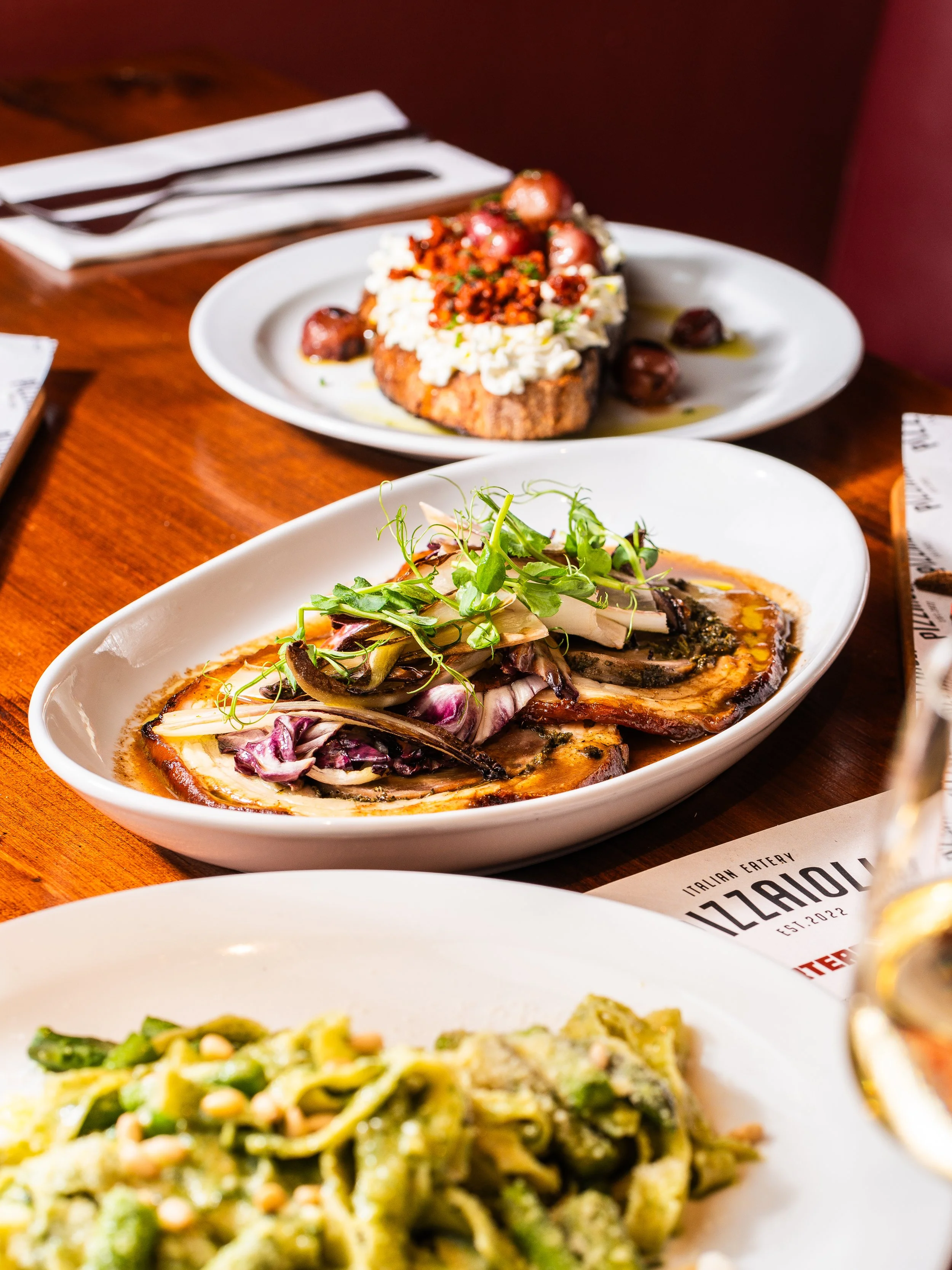 Three dishes of Italian food on a wooden table: grilled vegetables with microgreens, eggplant with garnish, and pasta with green sauce.