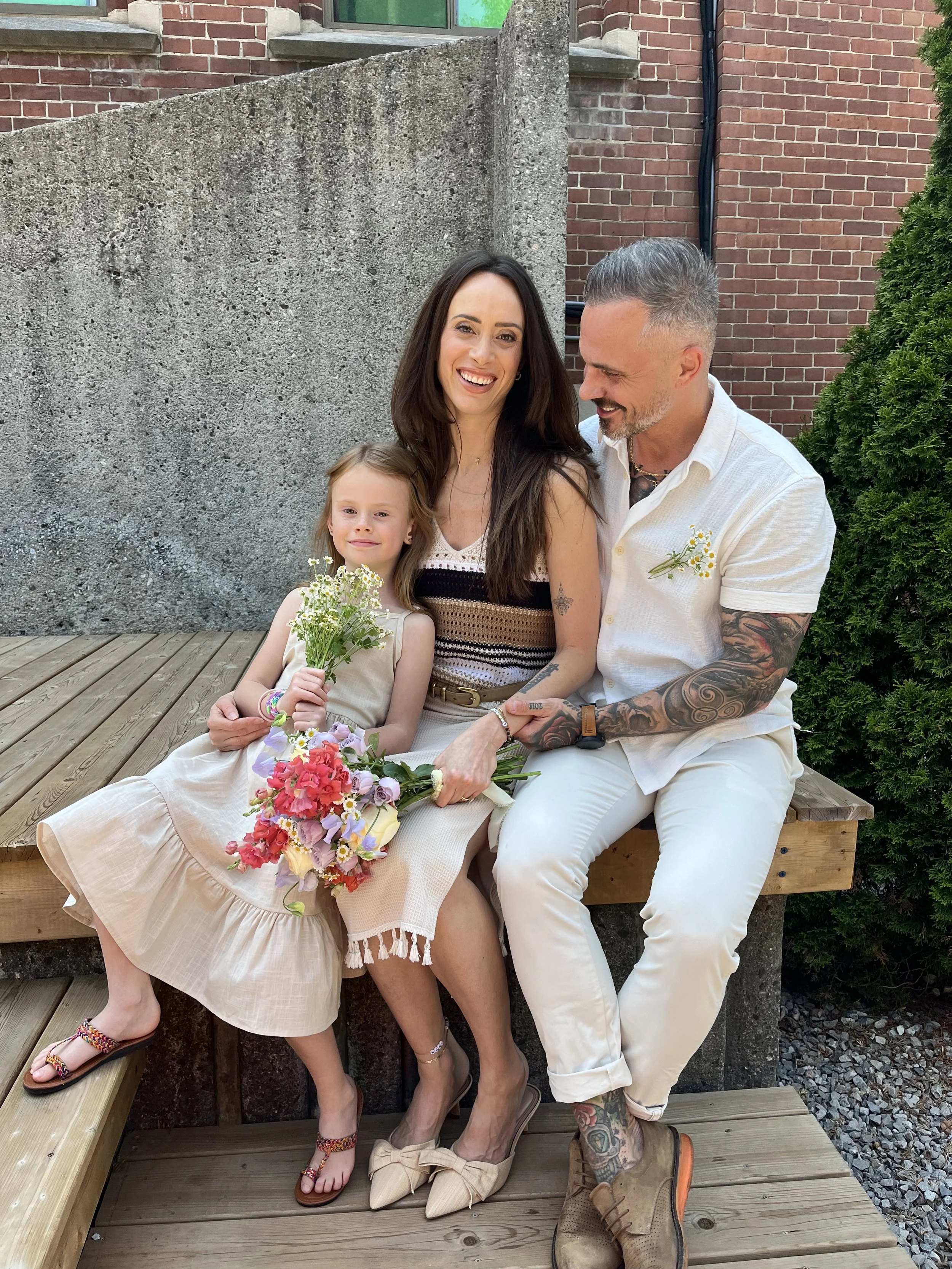 A family of three sitting on a wooden bench outdoors. The woman with long dark hair, wearing a striped dress, is smiling at the camera. The young girl in a beige dress holding a bouquet of flowers also looks at the camera. The man with tattoos, short gray hair, wearing a white shirt and beige pants, is smiling and looking at the woman.