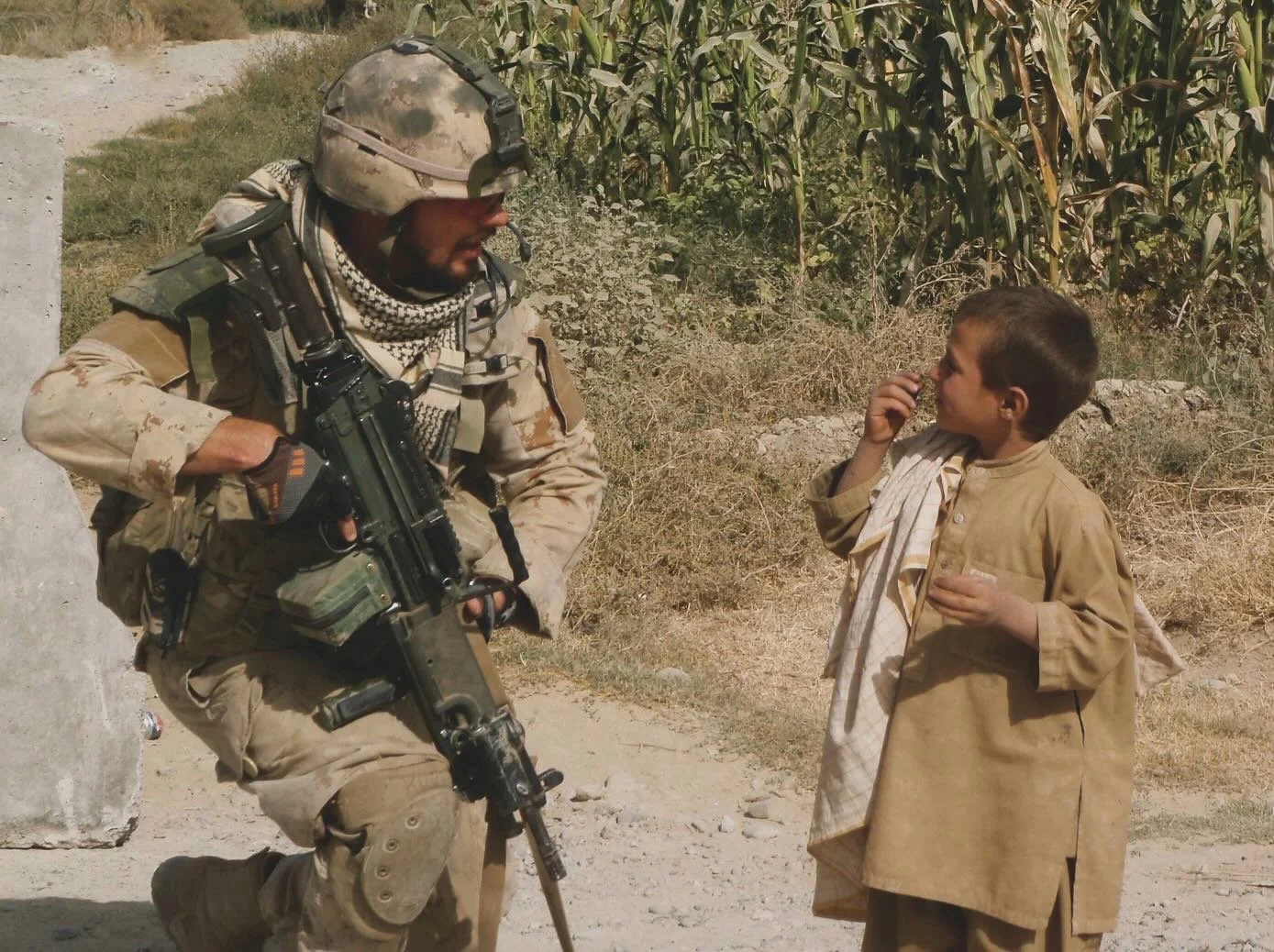 A soldier in camouflage gear and helmet engaged in conversation with a young boy in traditional clothing, standing outdoors with cornfields in the background.
