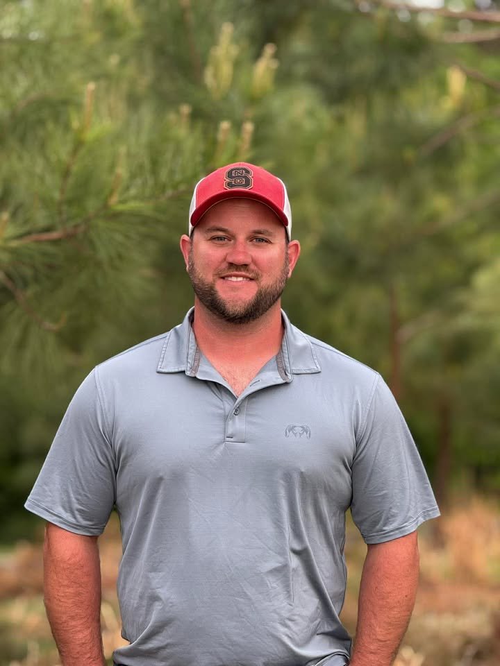 A man with a beard wearing a red baseball cap and a light gray polo shirt stands outdoors in front of green trees, smiling at the camera.