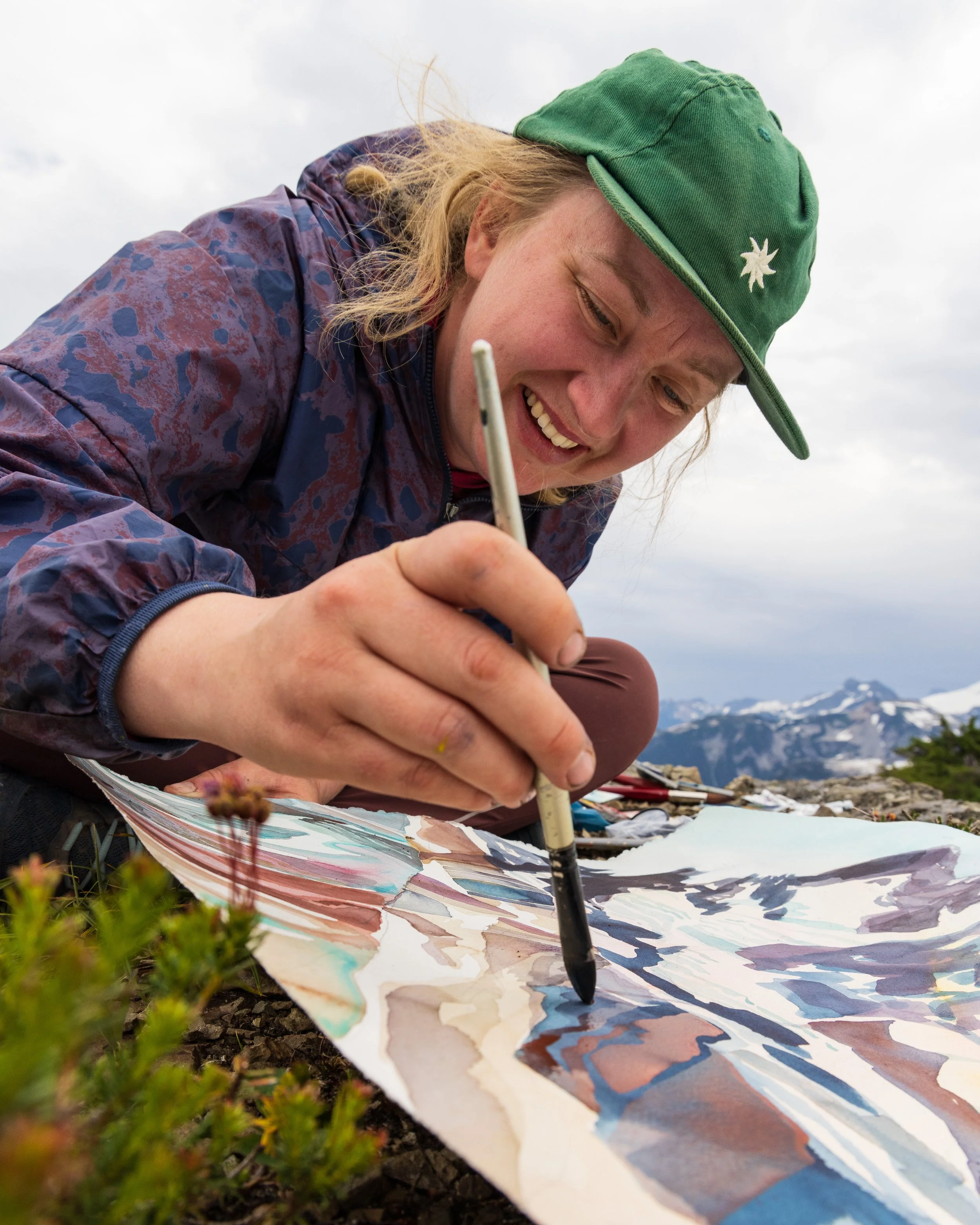 Woman drawing a landscape on paper outdoors with mountains in the background.