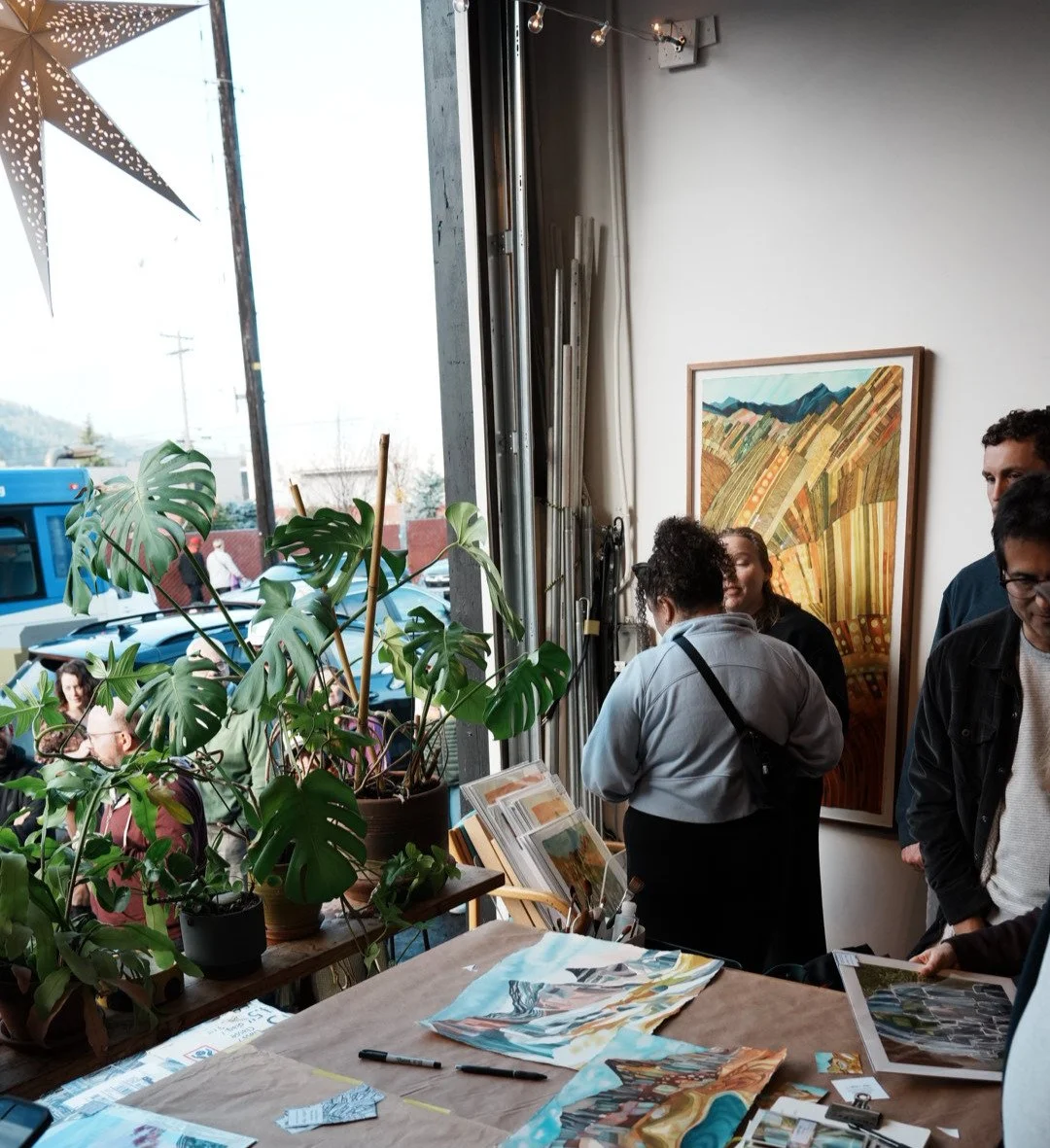People gather inside an art gallery with large windows, a colorful landscape painting on the wall, a table with art supplies, and potted plants near the window.
