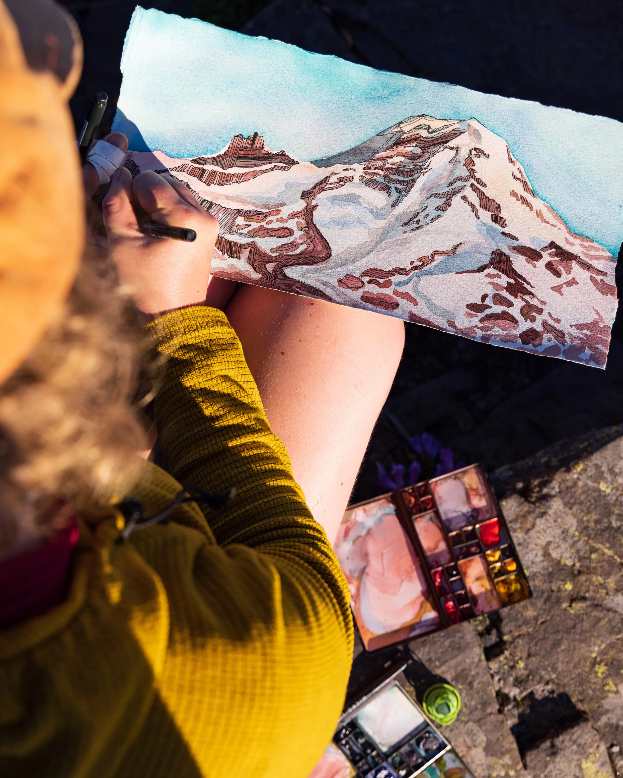 Person sitting outdoors, painting a landscape of snow-capped mountains and a winding path with watercolor paints.
