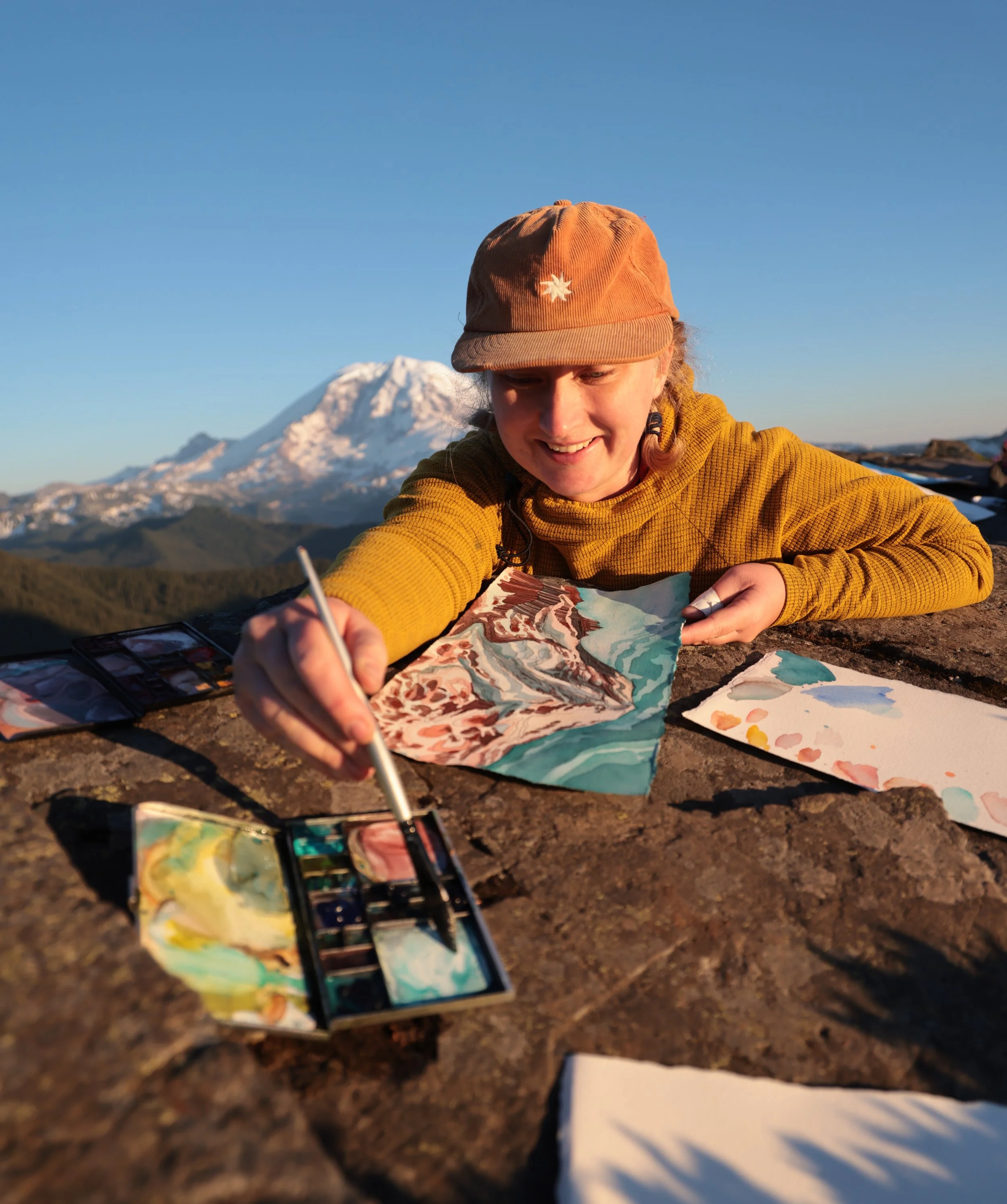 A woman painting outdoors on a stone surface with a mountain and snow-capped peak in the background, wearing a orange cap and a mustard yellow sweater.