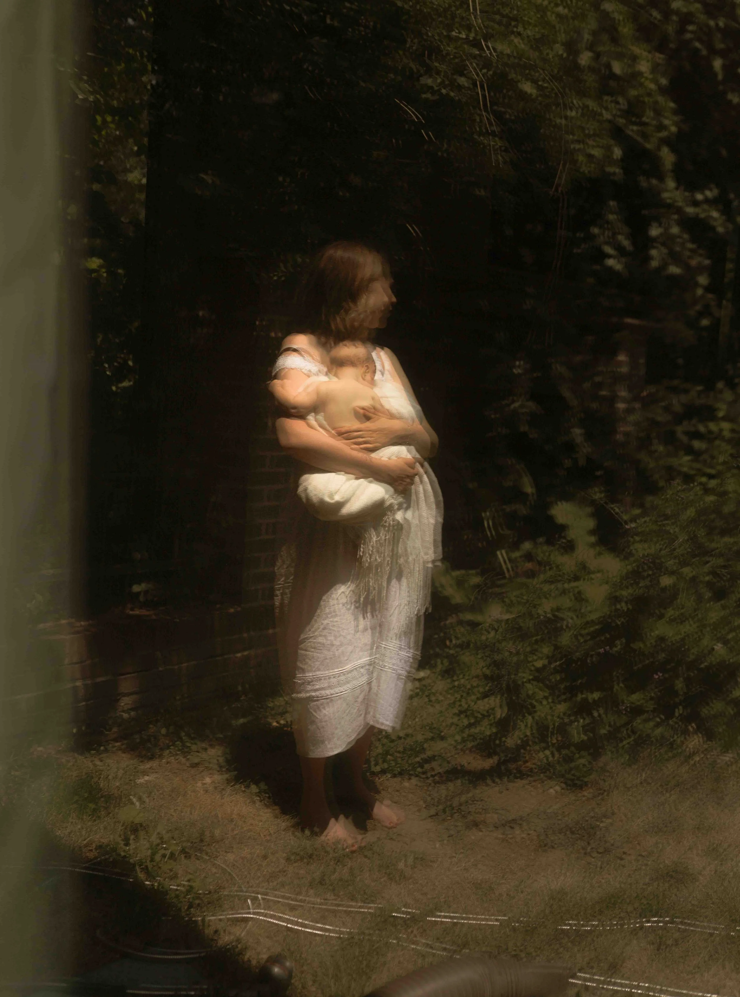 A woman wearing a white dress holding a baby, standing outdoors at night near a brick wall and trees.