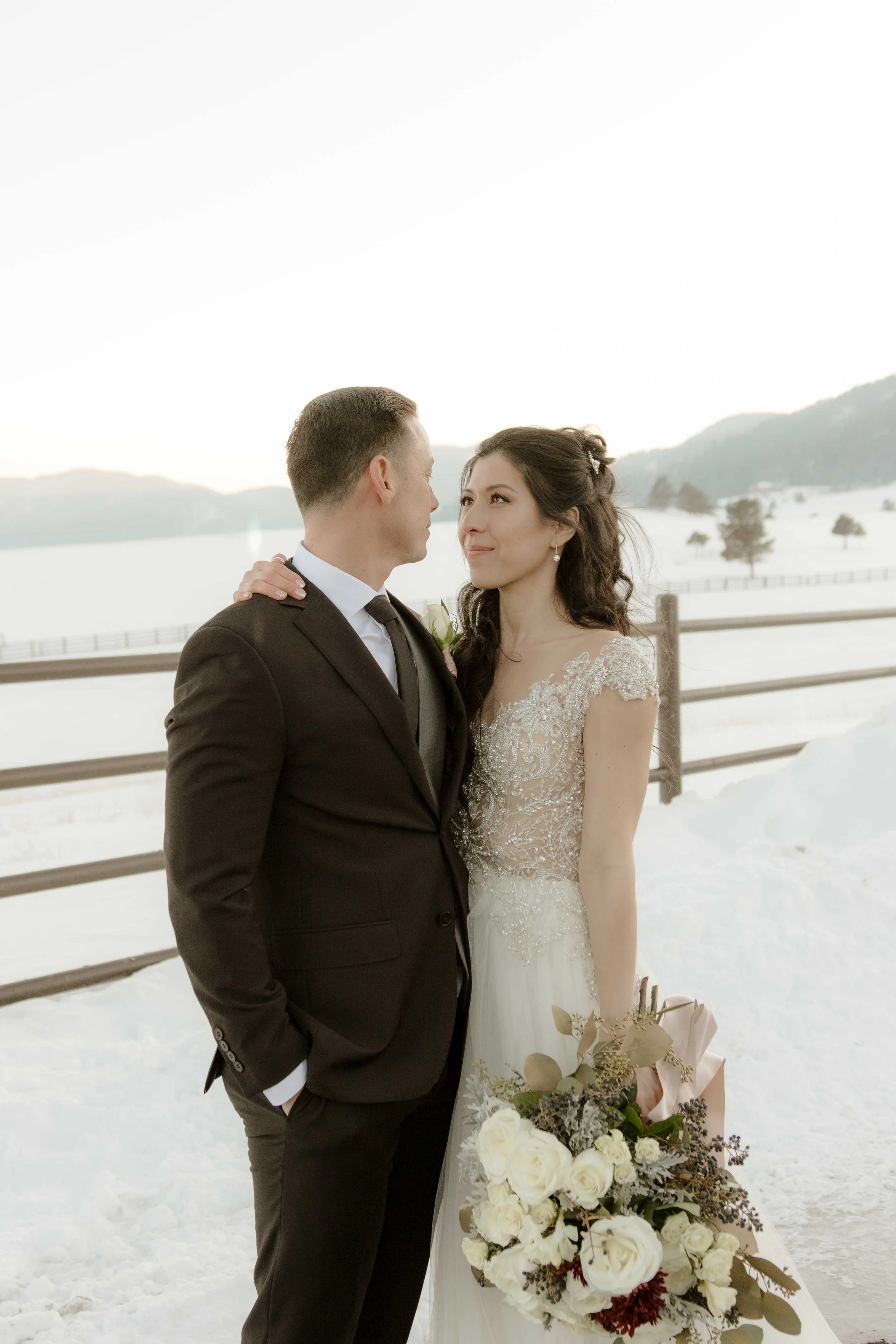 A bride and groom standing outdoors on snow, gazing into each other's eyes, with a lake and mountains in the background, during winter.