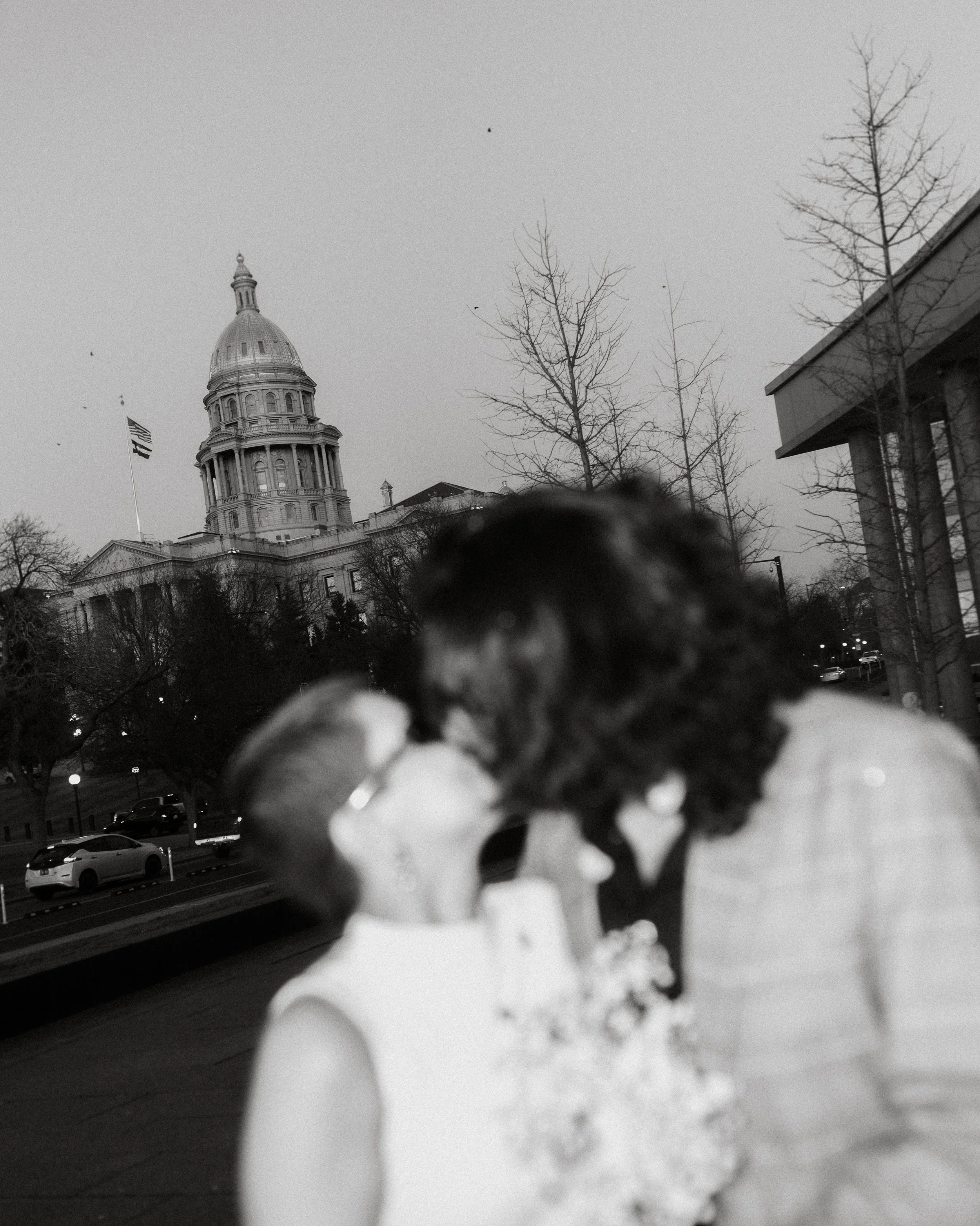 Blurred photo of a couple kissing outdoors with the Texas State Capitol building in the background.