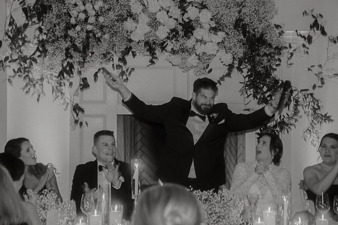 A man in a tuxedo is standing above a table, holding a phone in each hand with his arms outstretched, smiling. Four women and one man are seated at the table, clapping, smiling, and looking at him. The table has candles and glasses, with a floral arrangement hanging overhead.