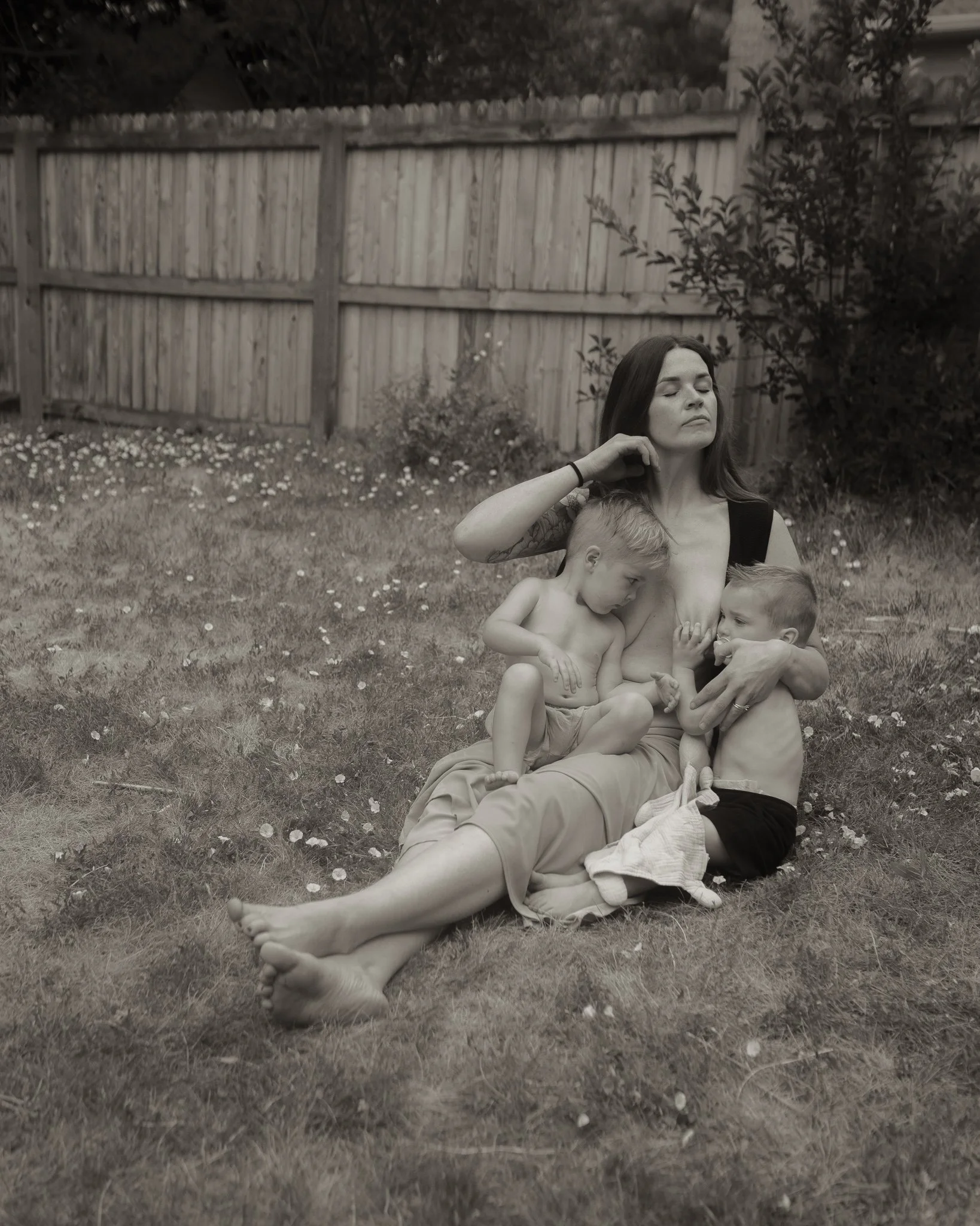 A woman with closed eyes sitting on the grass with two children, surrounded by flowers and trees, wooden fence in the background, black and white photo.