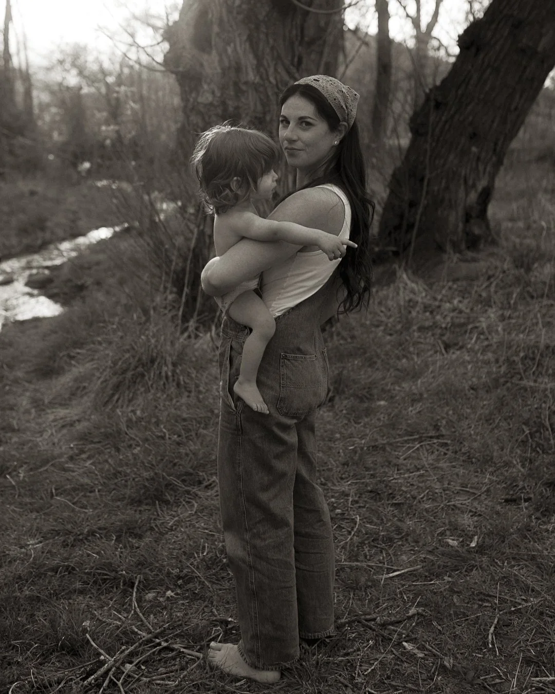 A woman holding a young child outdoors in a natural setting, with trees and a stream in the background, captured in black and white.