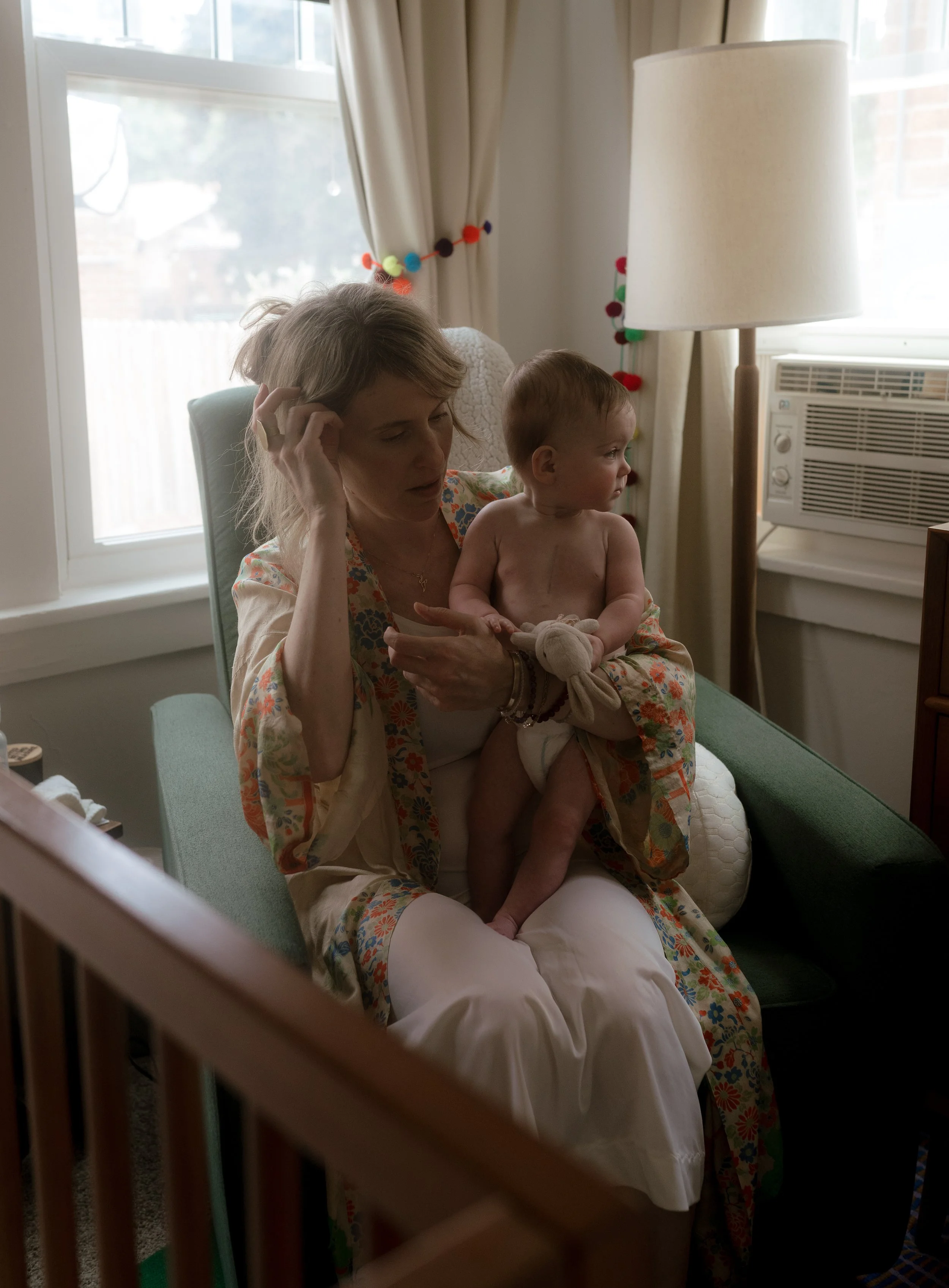 A woman sitting on a green armchair holding a shirtless baby in her lap, with a window and beige curtains in the background. The woman appears to be brushing her hair.