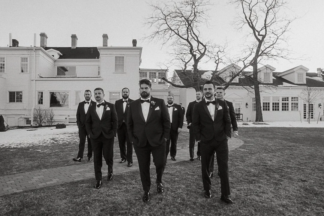 A group of men in tuxedos walking outdoors on a yard with a large white house in the background, leafless trees, and patches of snow.