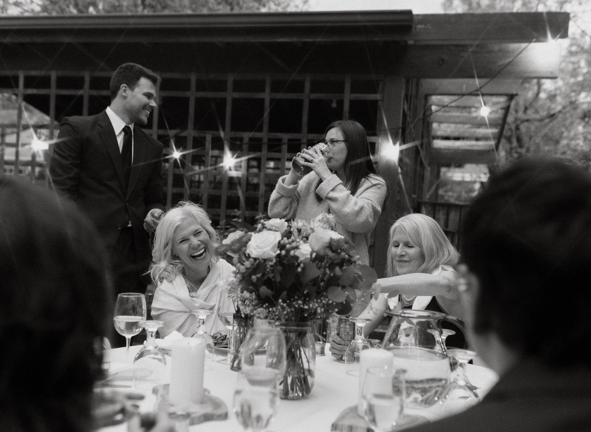 two women laugh and people talk around a table at a backyard wedding reception