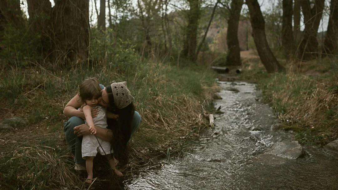 A woman and a small girl hugging by a creek in a wooded area.