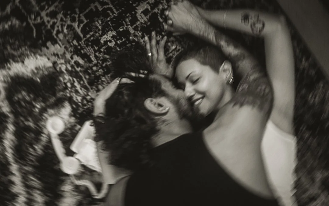 Two women lying on a patterned blanket, facing each other, smiling and touching their foreheads together.