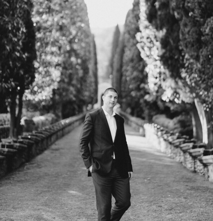 A man wearing a suit standing on a pathway lined with tall, trimmed trees.