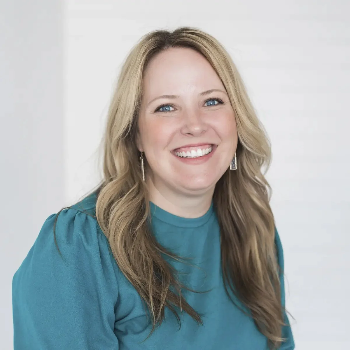 A smiling woman with long wavy blonde hair, blue eyes, wearing a teal top and silver earrings, standing against a plain light background.