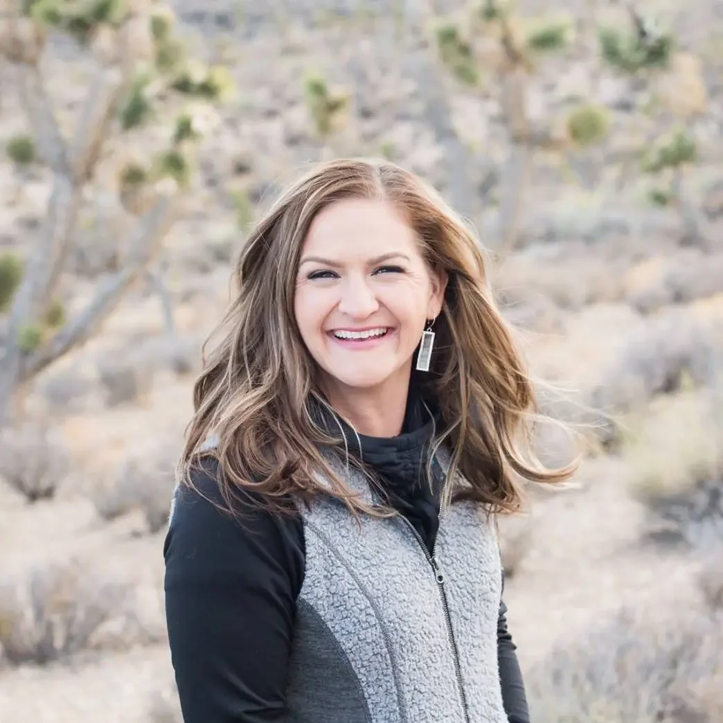 A woman with long brown hair smiling outdoors with desert vegetation in the background.