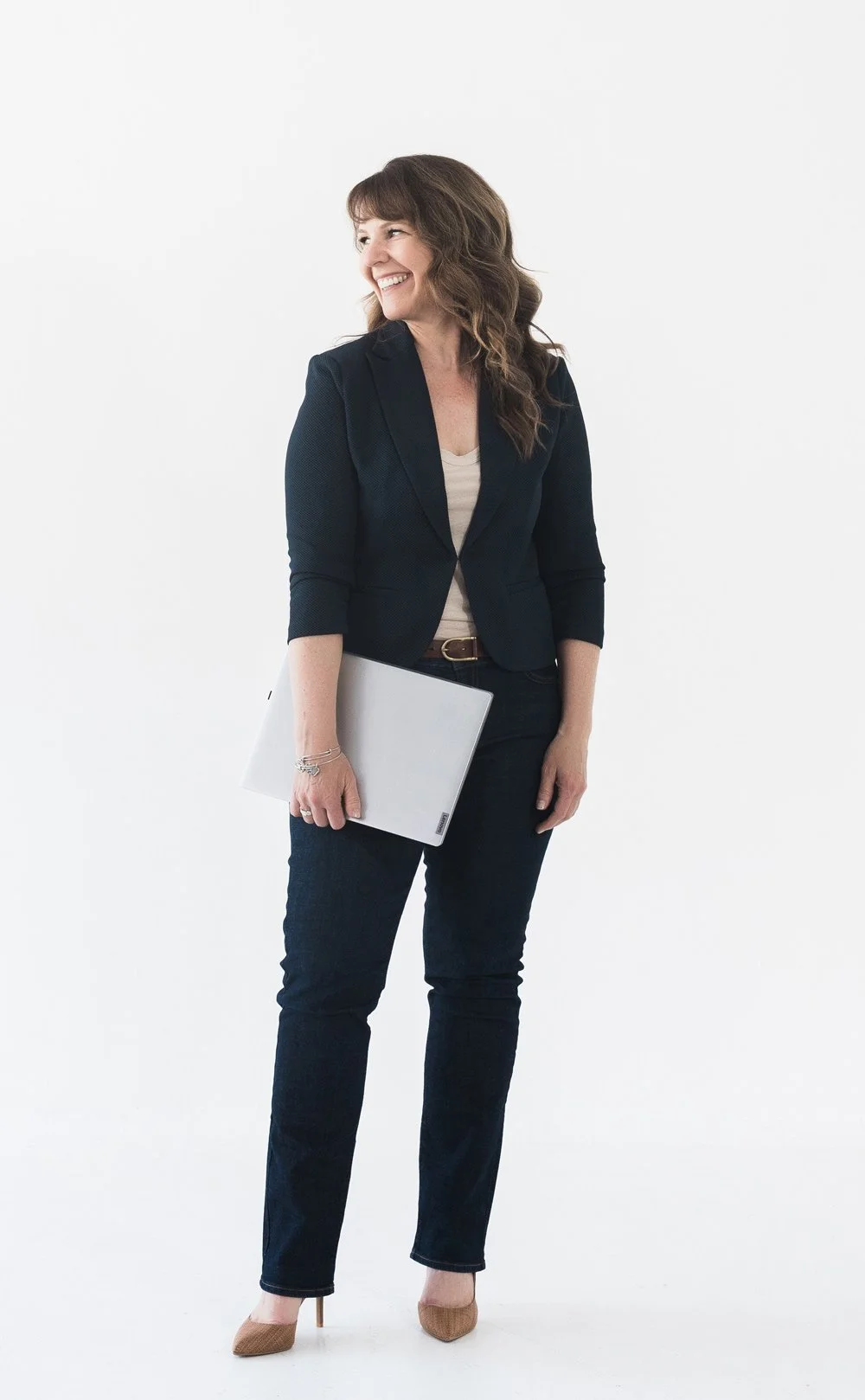 A woman with brown wavy hair wearing a black blazer, light beige top, black pants, and beige high heels holding a silver laptop, standing against a white background, smiling and looking to the side.