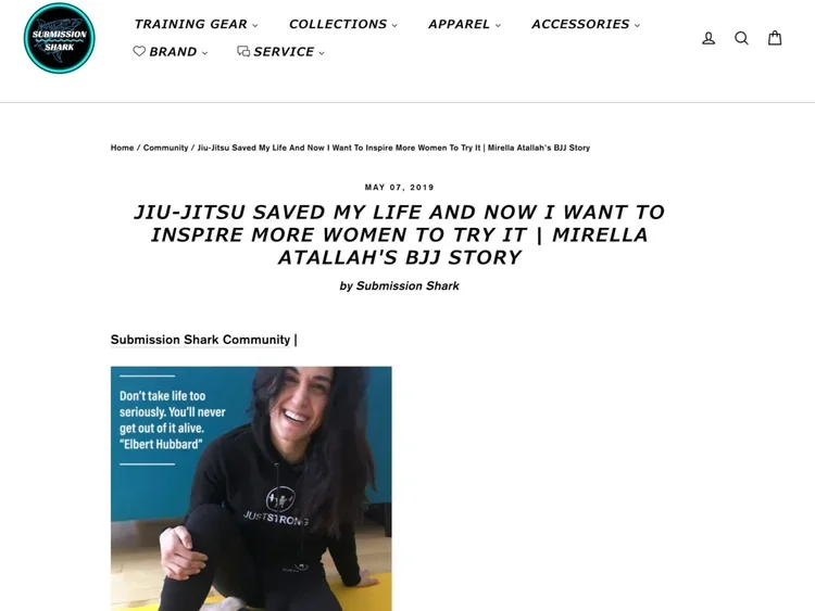 A woman with long dark hair smiling, wearing a black hoodie with a logo, sitting on the floor in a martial arts or jiu-jitsu training environment.