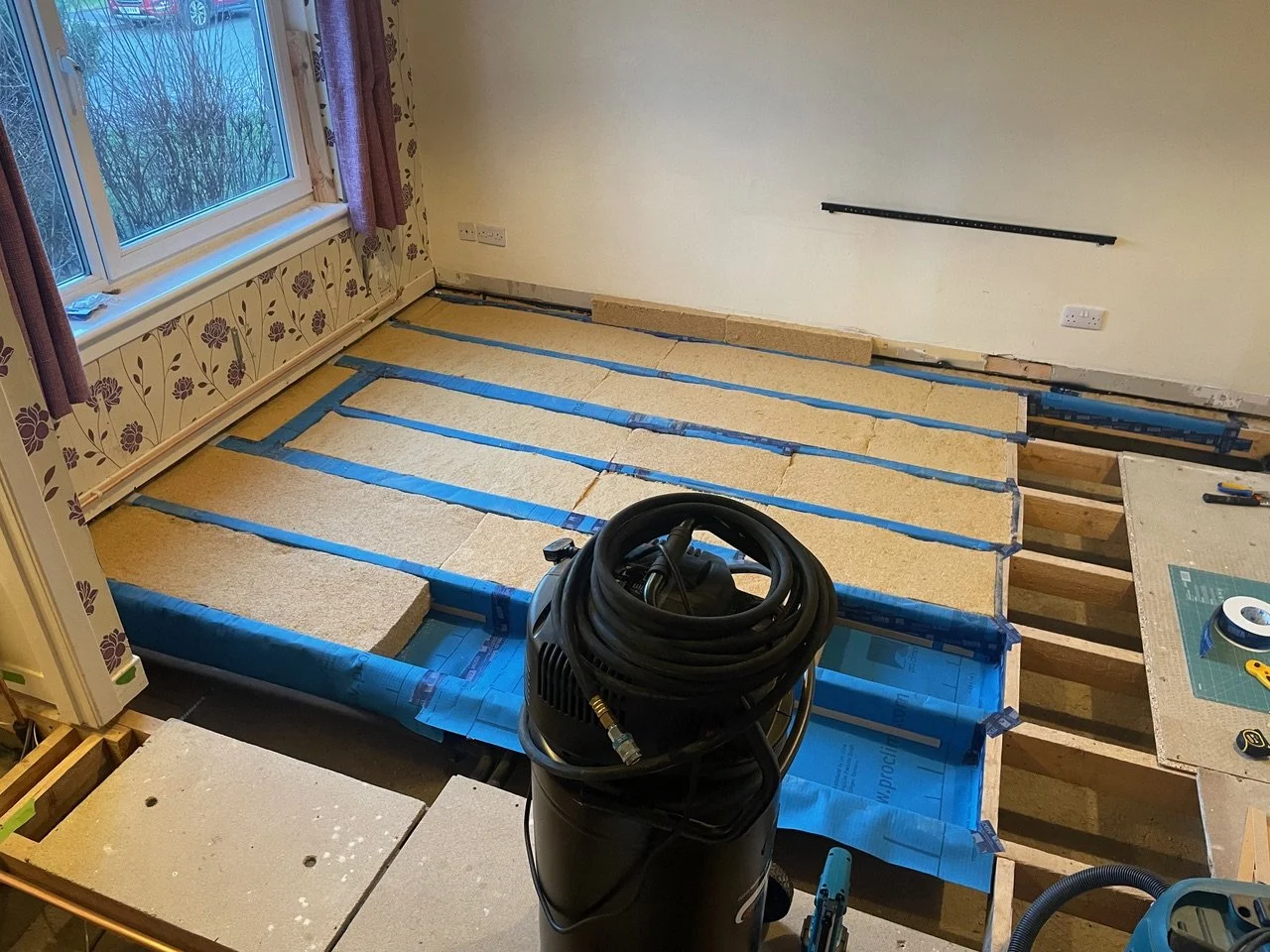 Room under renovation with flooring removed, showing wooden subfloor and support beams, vacuum cleaner in the center, window with floral curtains on the left, and construction tools on the right.