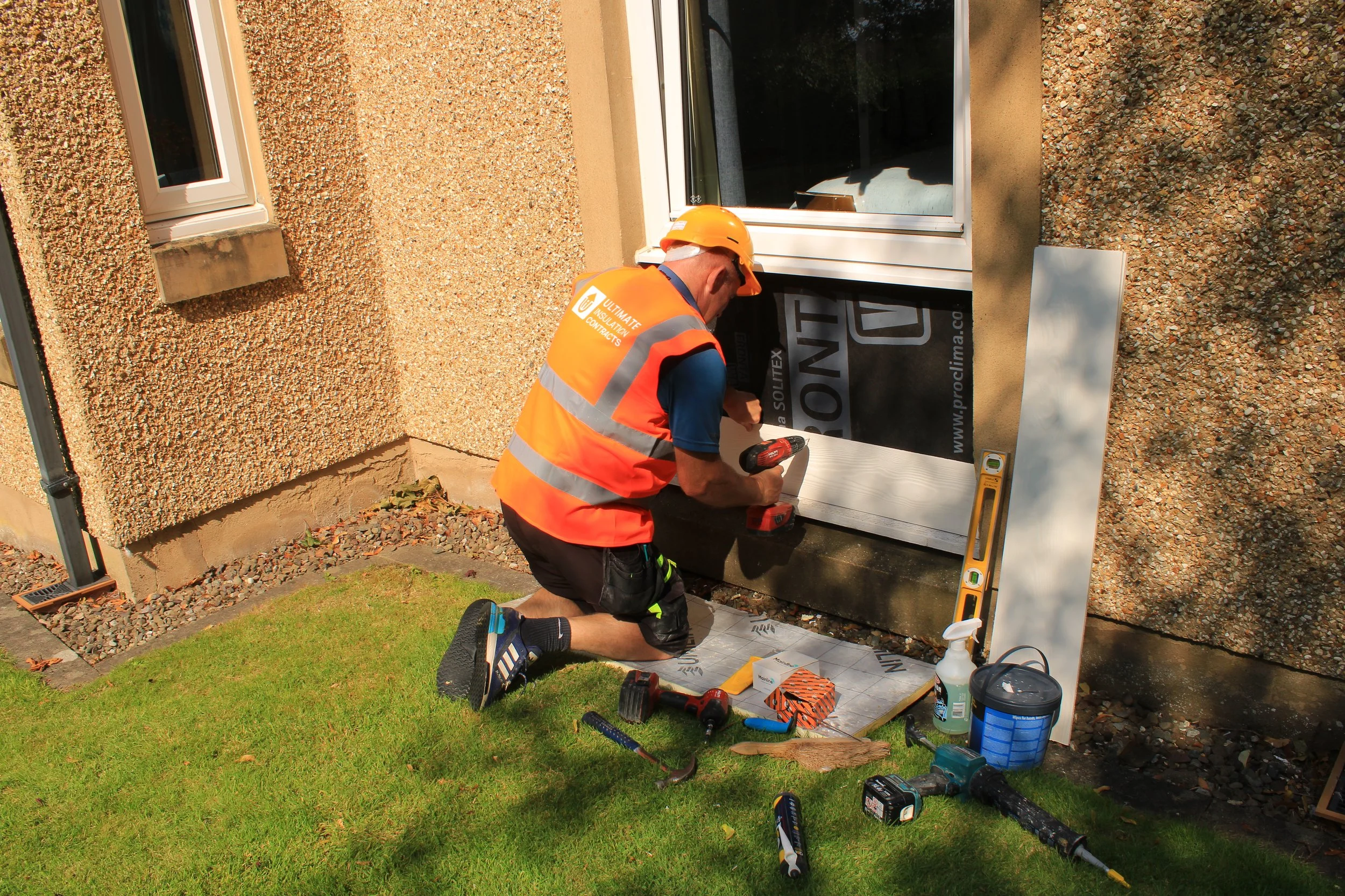 A construction worker kneels on the grass installing a window at a house. He wears an orange safety vest, an orange hard hat, and uses power tools and other construction materials around him.