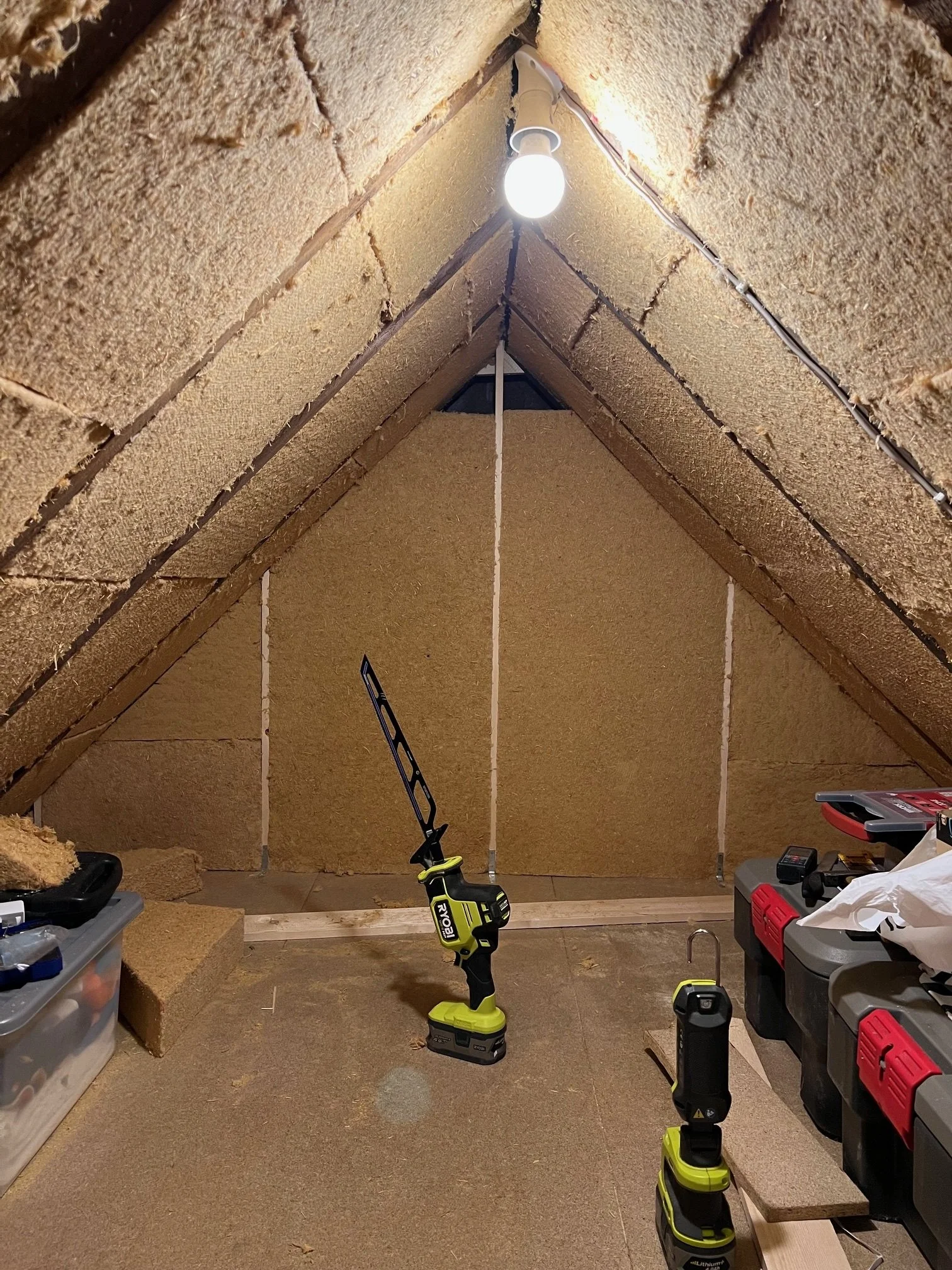Attic space under construction or renovation with insulation on the walls and ceiling. There are various tools including a cordless drill, flashlight, and storage bins.
