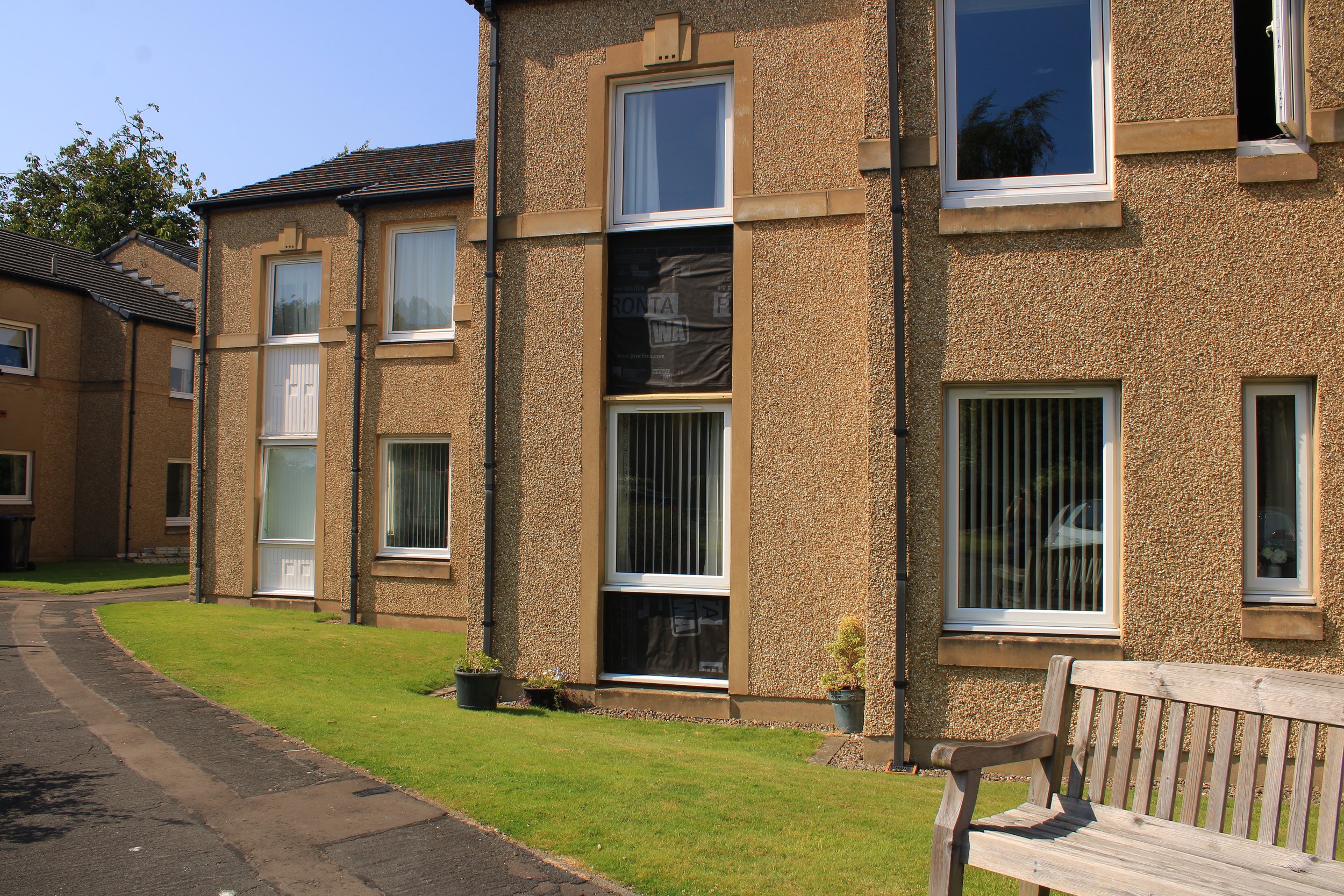 Exterior view of apartment building with four windows, one door, a small potted plant, and a wooden park bench on a grassy area. Clear blue sky in the background.