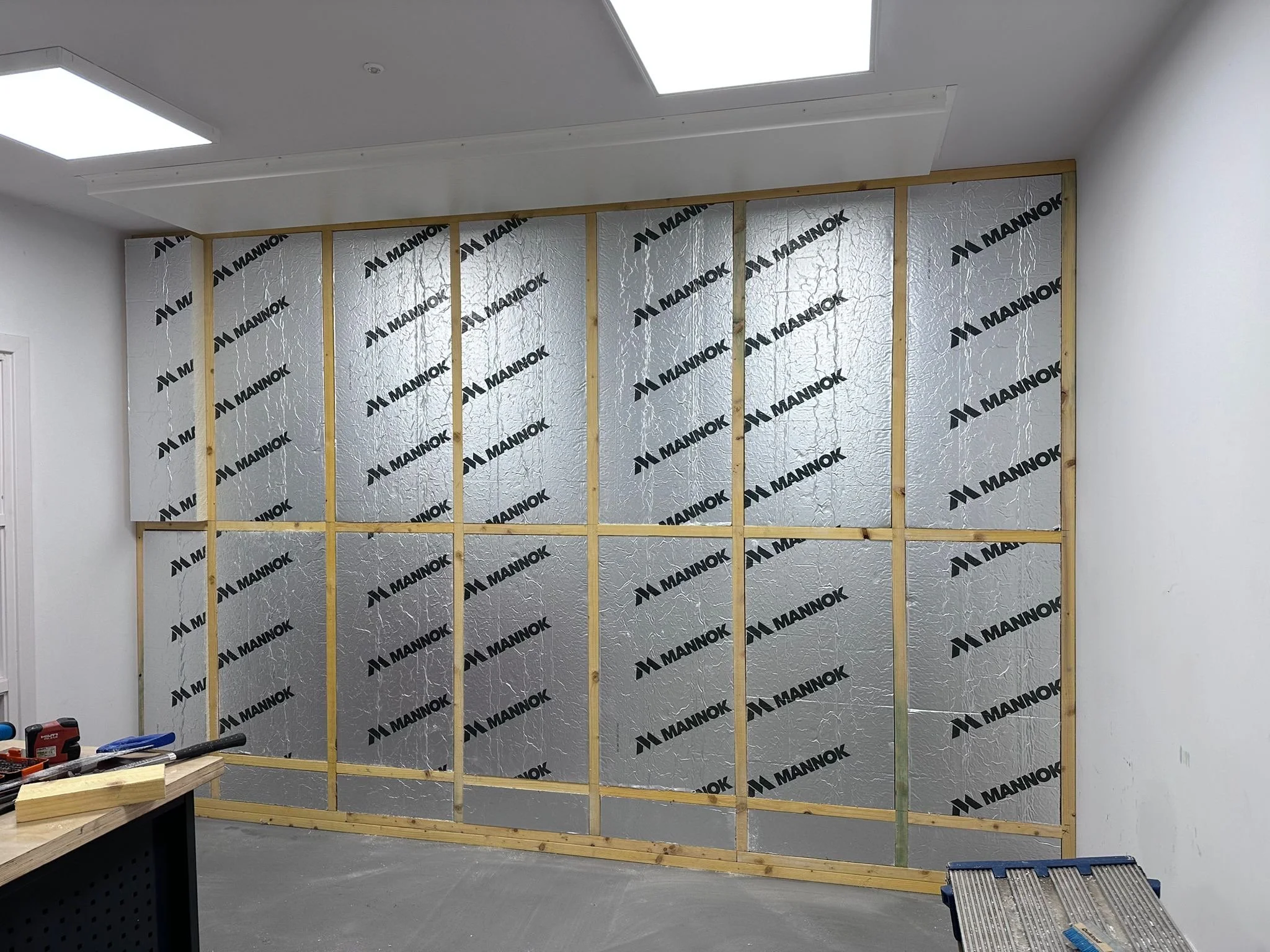 Interior of a room with a partially constructed wall, insulation panels with the brand name 'MANNOK,' tools and building materials on a workbench, and a light-colored ceiling.