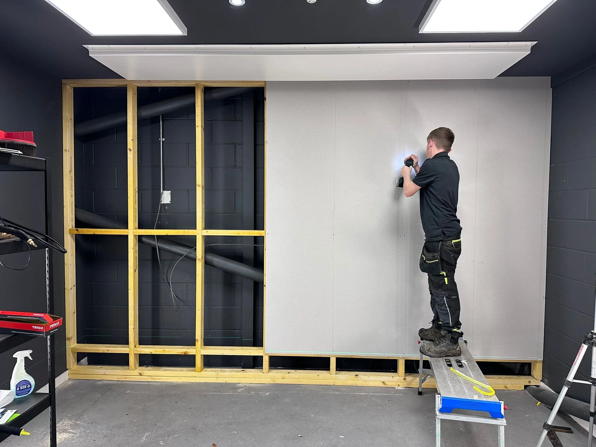 A man in black work clothes standing on a ladder, working on a wall with partial wooden framing and drywall. Construction tools and materials are nearby.
