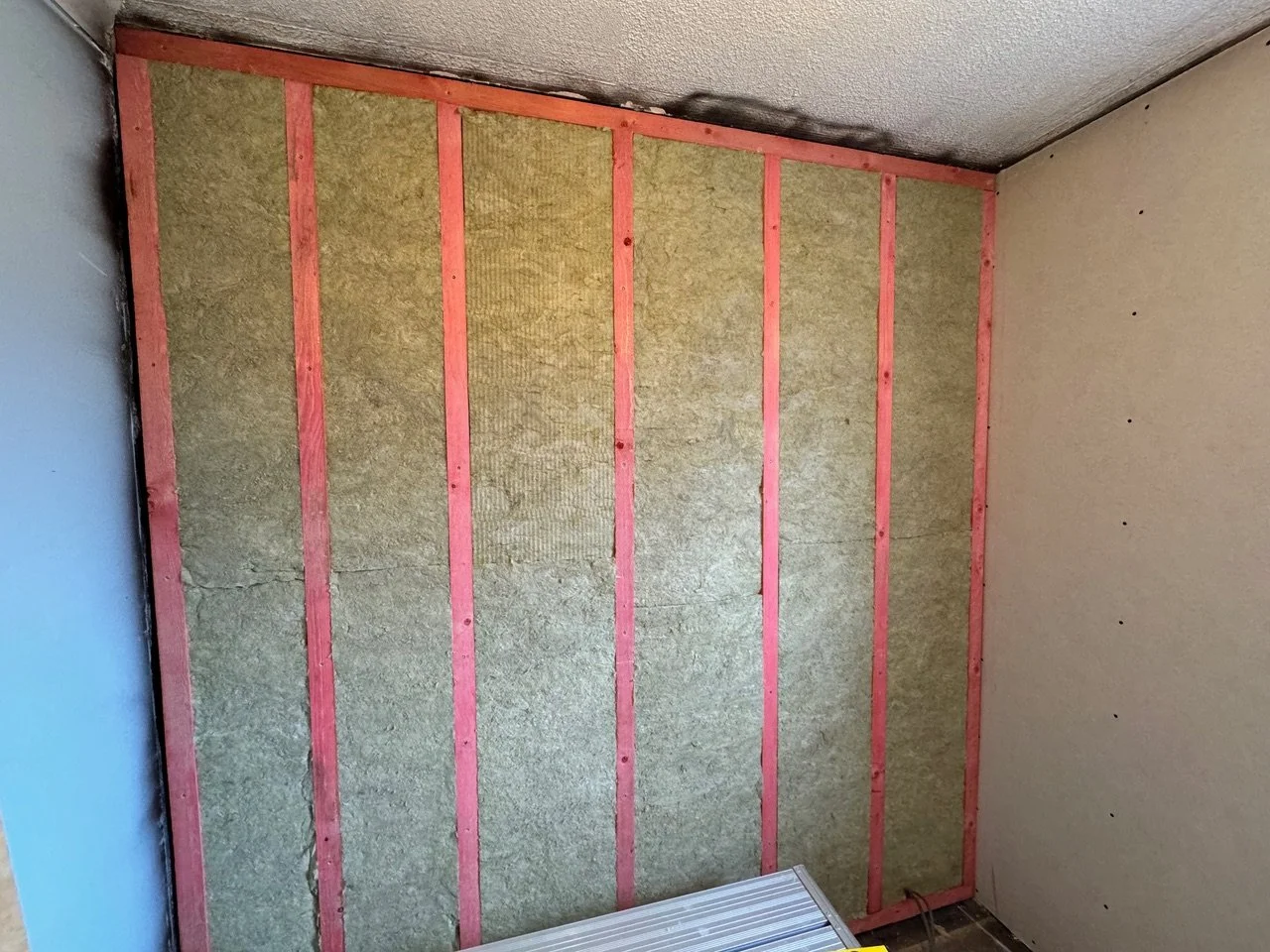 Interior wall undergoing renovation with insulation installed between pink wooden studs, and drywall partially attached to the right side.