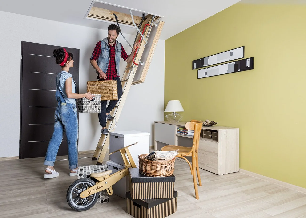 Two people moving belongings into an apartment with a staircase attic hatch, with boxes and a scooter nearby.
