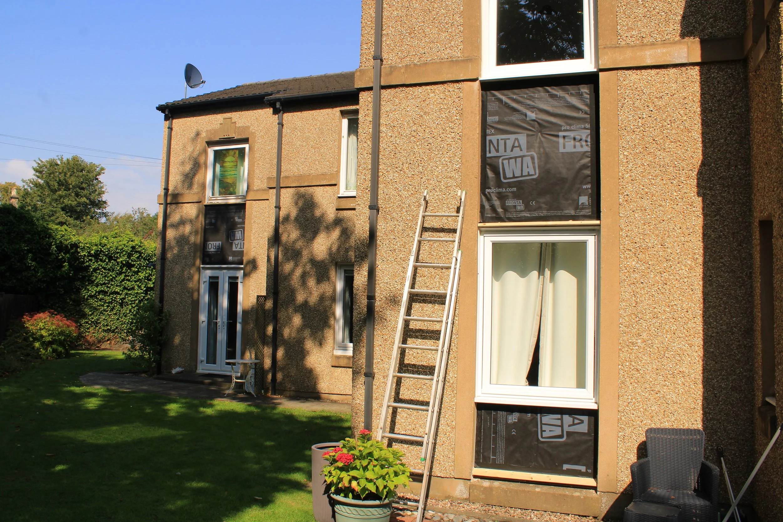 A two-story house with a textured exterior wall under renovation, showing black weather-resistant sheathing on some windows, a ladder leaning against the wall, and a satellite dish on the roof. The yard has green grass, a tree, and potted plants.