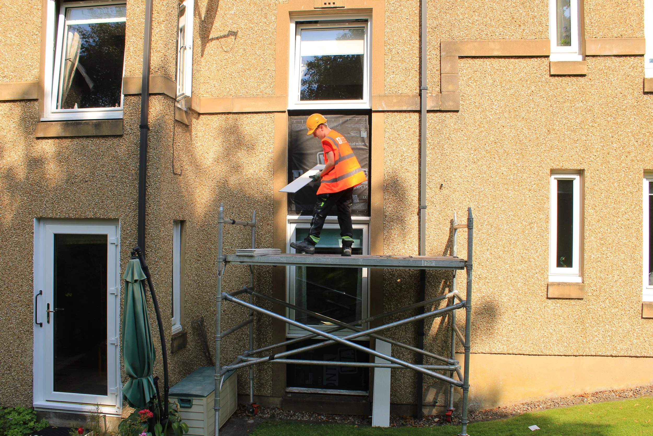 A construction worker wearing an orange safety vest and helmet is working outside a beige brick house on a scaffold, installing or repairing a window.