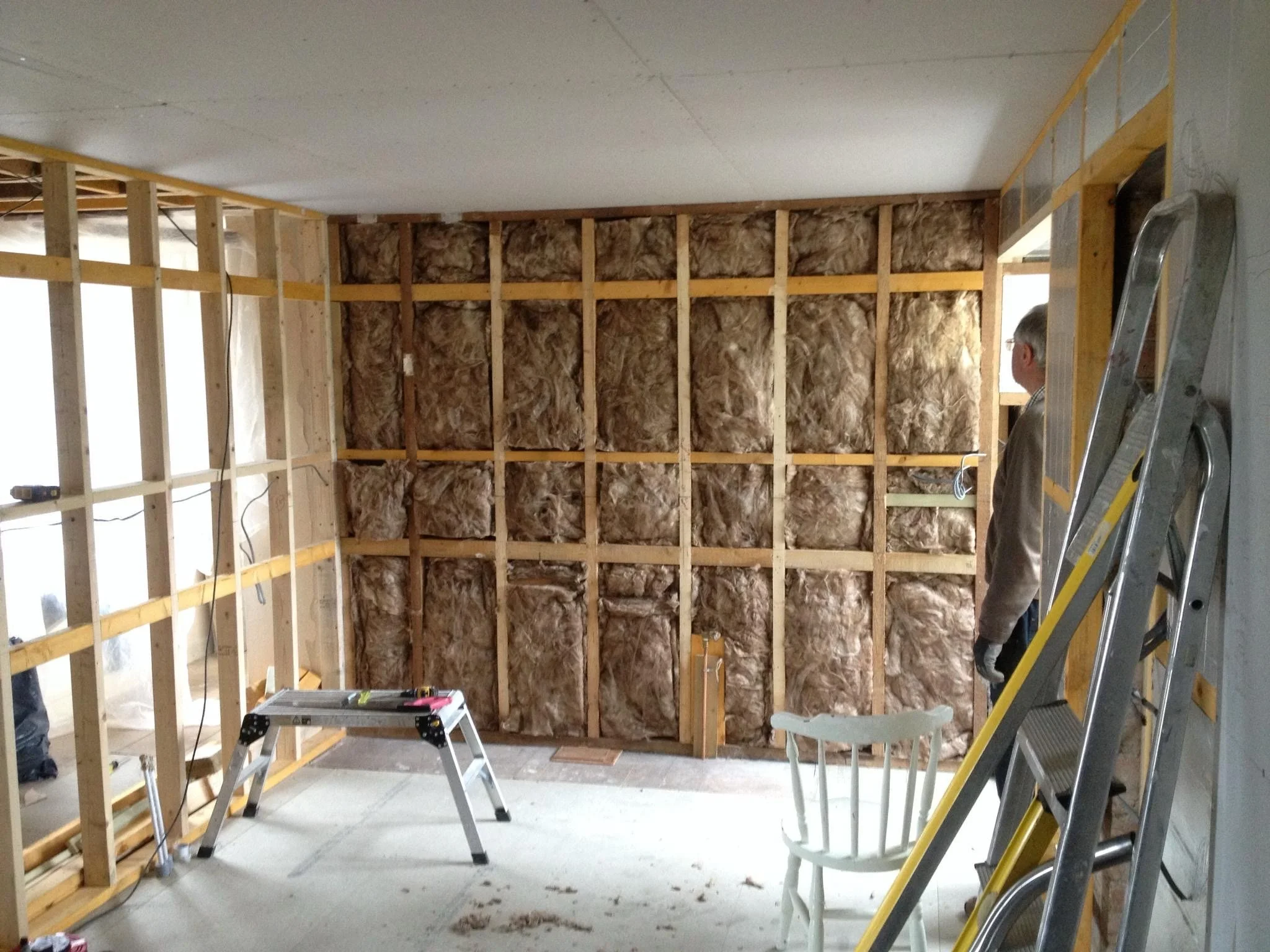 A partially constructed interior wall with wooden framing and insulation, with ladders and construction tools visible, and a person in the background.