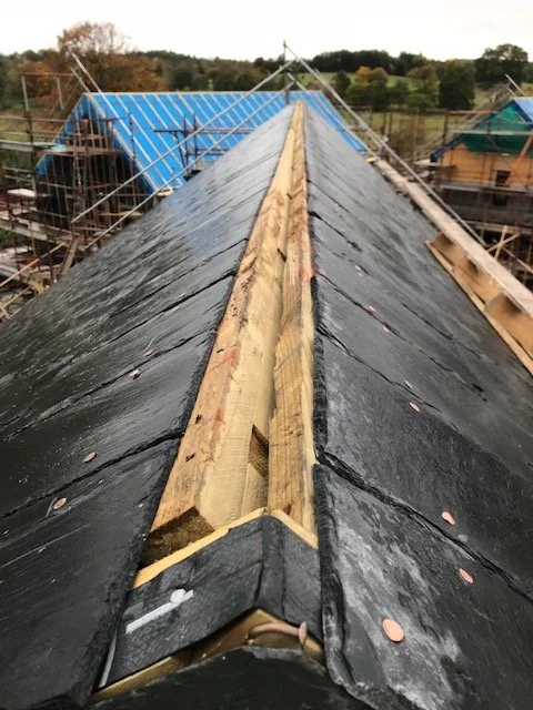 Roof under construction with wooden support beam and black roofing material, scaffolding and other buildings in background.
