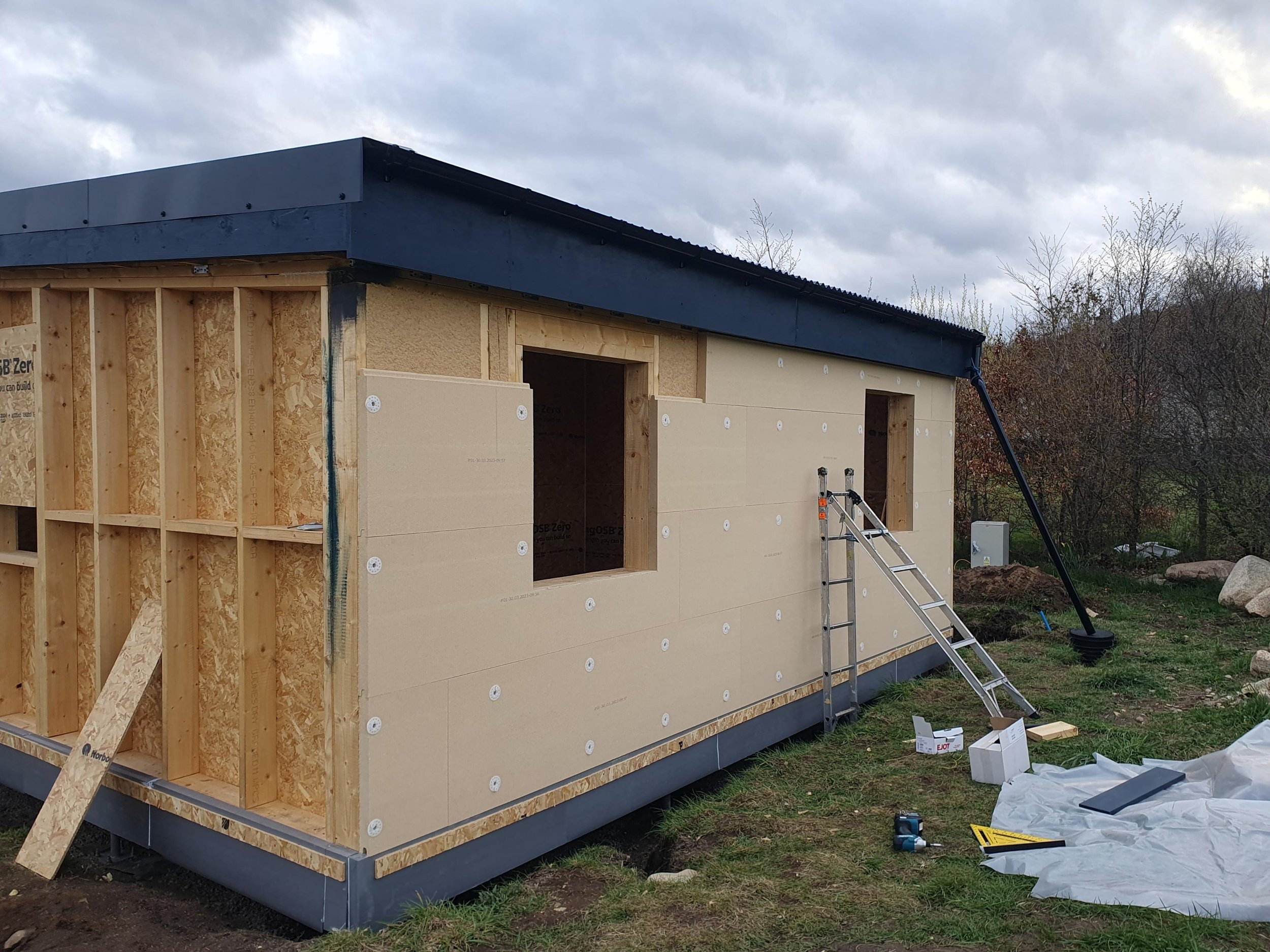 Construction of a small building or house with wooden framing and exterior wall insulation panels being installed. Tools, a ladder, and building materials are visible on the ground outside. The sky is overcast, and trees are in the background.